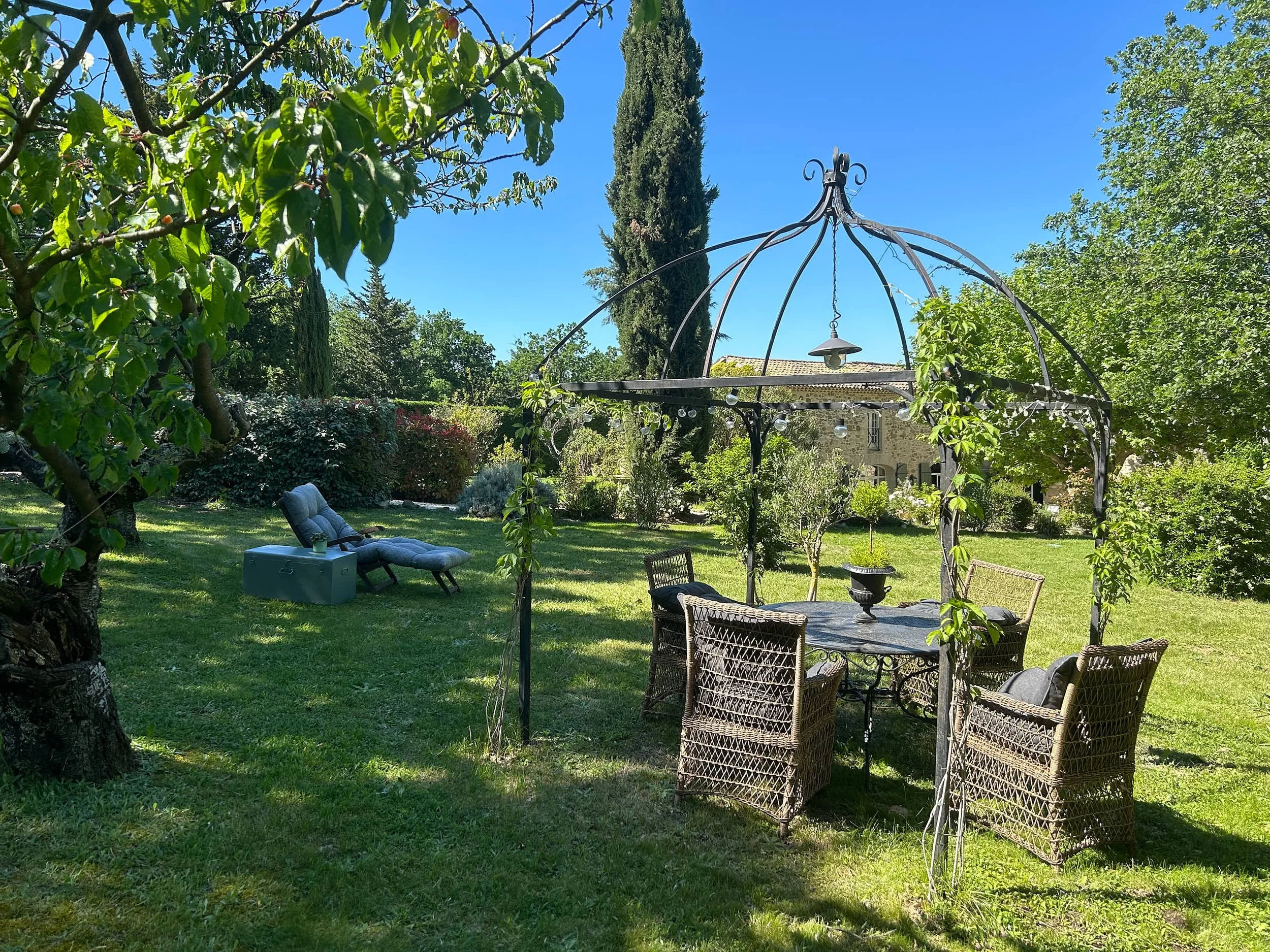 Garten mit einem Metallpavillon, einem Rattan-Esstisch mit Stühlen, einem Liegestuhl mit Auflage, umgeben von Bäumen und Sträuchern, im sonnigen Himmel.
