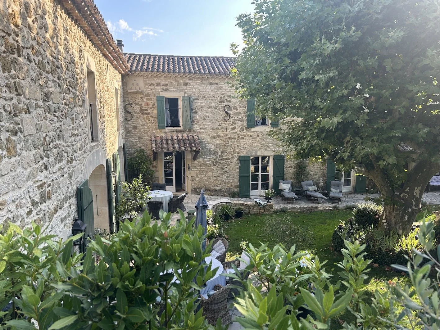 Ein Innenhof mit Steingebäuden, grünen Fensterläden, einer großen Kastanie, einem Sitzbereich mit Liegestühlen und einer Sitzecke mit einem Esstisch. Sonnenschein und blauer Himmel sichtbar.