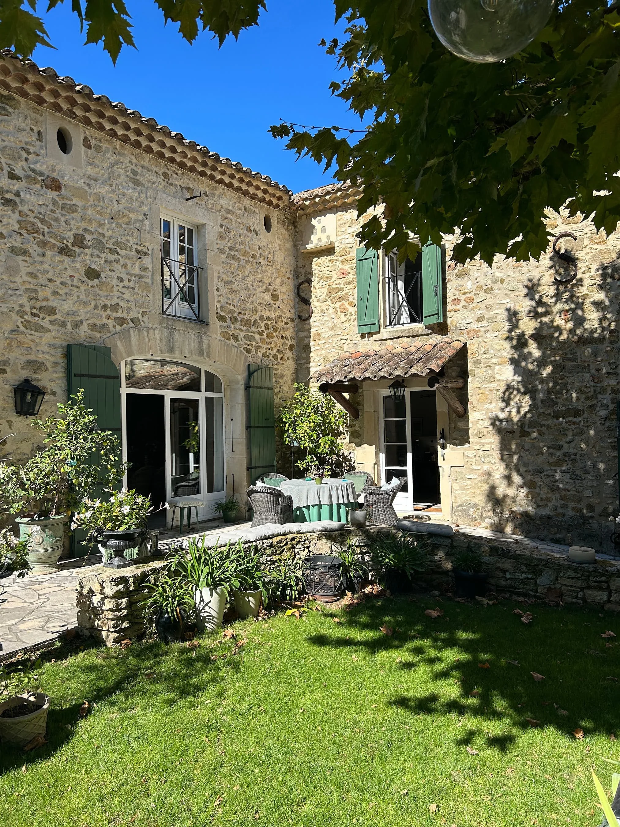 Ein gemütlicher Gartenbereich mit Steinterrasse, auf dem sich ein runder Tisch mit grüner Tischdecke und verschiedene Gartenmöbel befinden. Das Haus ist aus Stein mit grün gestrichenen Fensterläden, umgeben von Pflanzen und Bäumen bei sonnigem Himmel