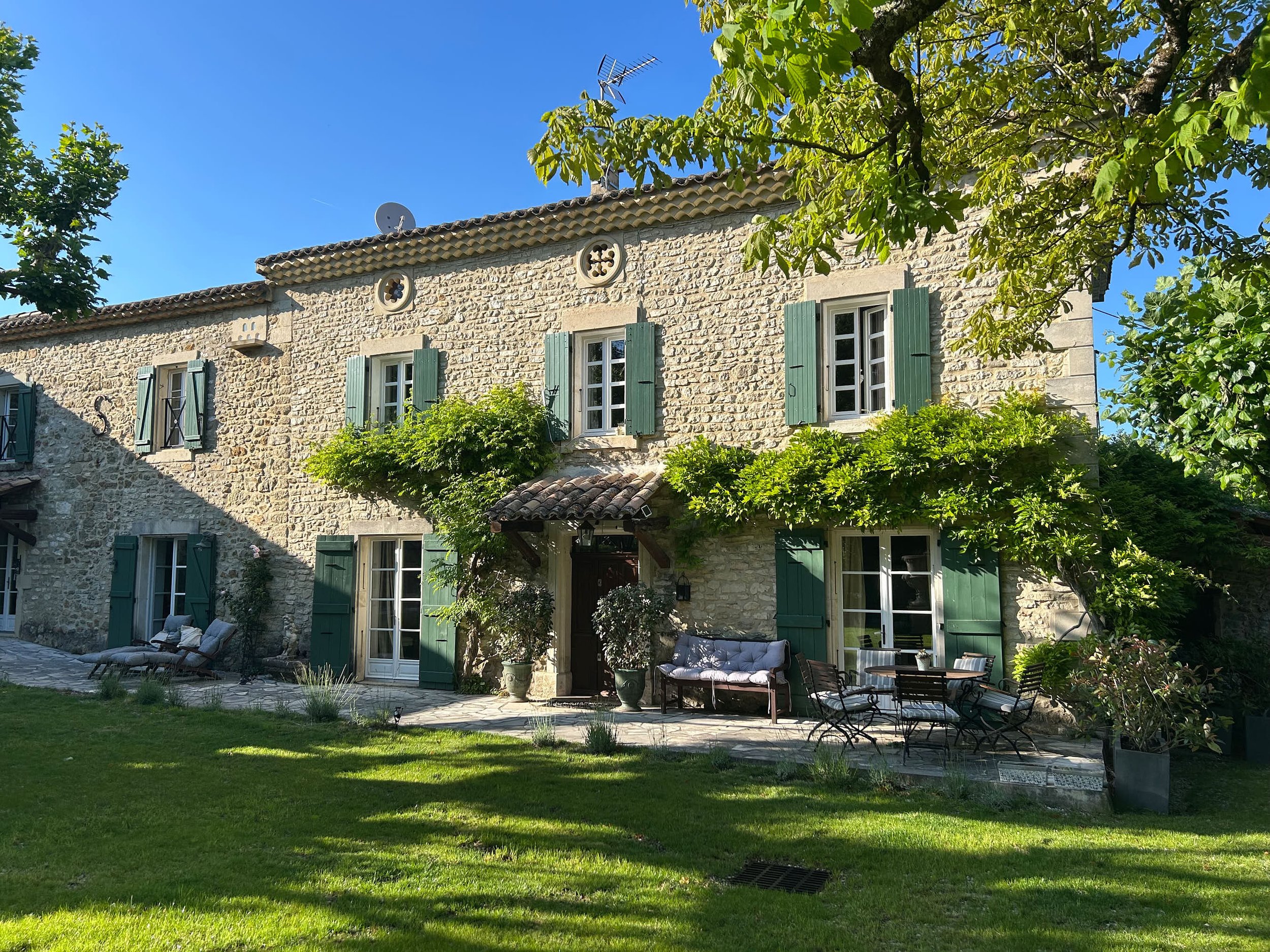 Ein altes Steinhaus mit grünen Fensterläden, umgeben von Bäumen und einem gepflegten Garten mit Gartenmöbeln im Freien, bei schönem Wetter.