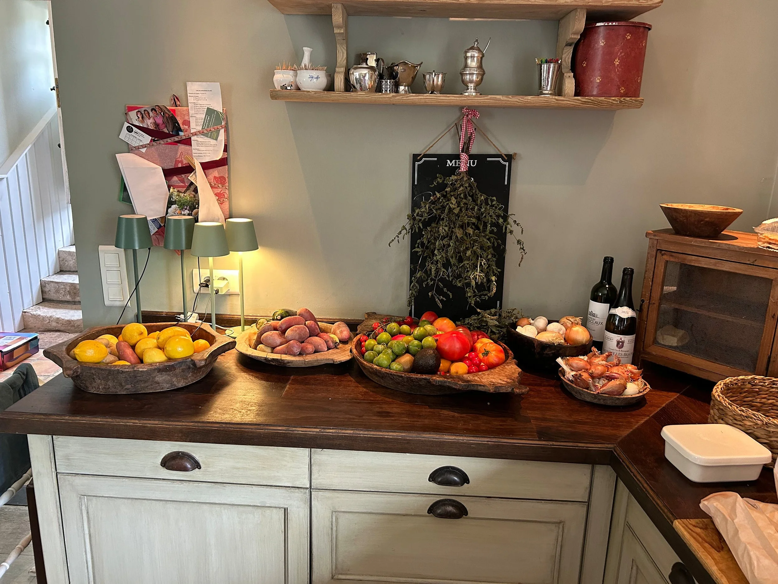 Küche mit Holztisch, auf dem verschiedene Obst- und Gemüsesorten in Holzschalen liegen, sowie Weinflaschen und eine kleine Glasvitrine. Im Hintergrund ist eine Wand mit einer Schatztruhe und einer Pinnwand mit Papieren und Fotos zu sehen.