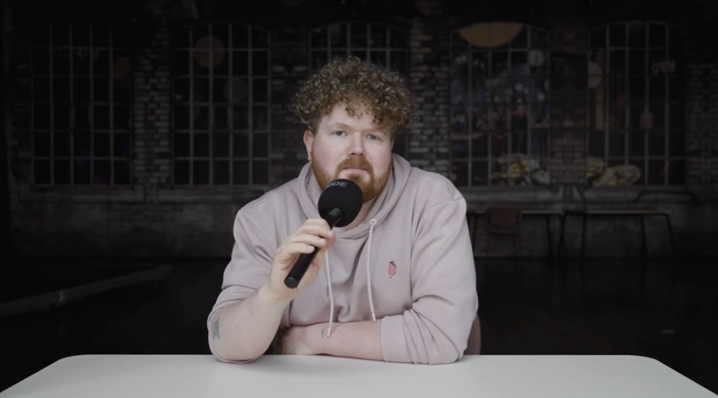 Kolja Fach sitzt an einem Tisch, hält ein Mikrofon und schaut direkt in die Kamera, im Hintergrund eine dunkle Backsteinwand.