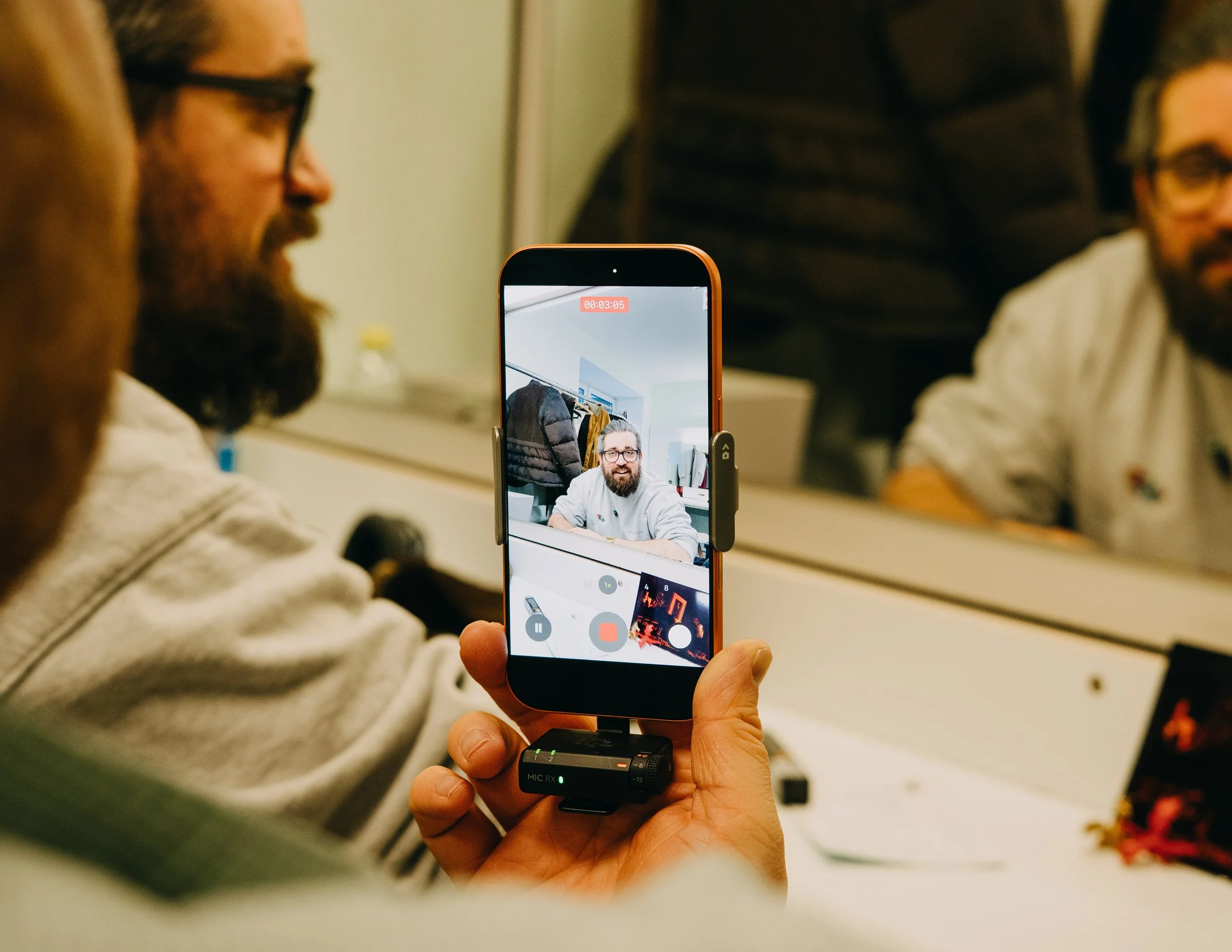 Nahaufnahme einer Hand, die ein Smartphone auf einem Tisch hält. Das Smartphone zeigt ein Video mit einem Mann, der eine weiße Jacke trägt, vor einem Hintergrund mit Kleidung. Auf dem Bildschirm sind Videoaufnahmetimer und Aufnahme-Icons sichtbar.