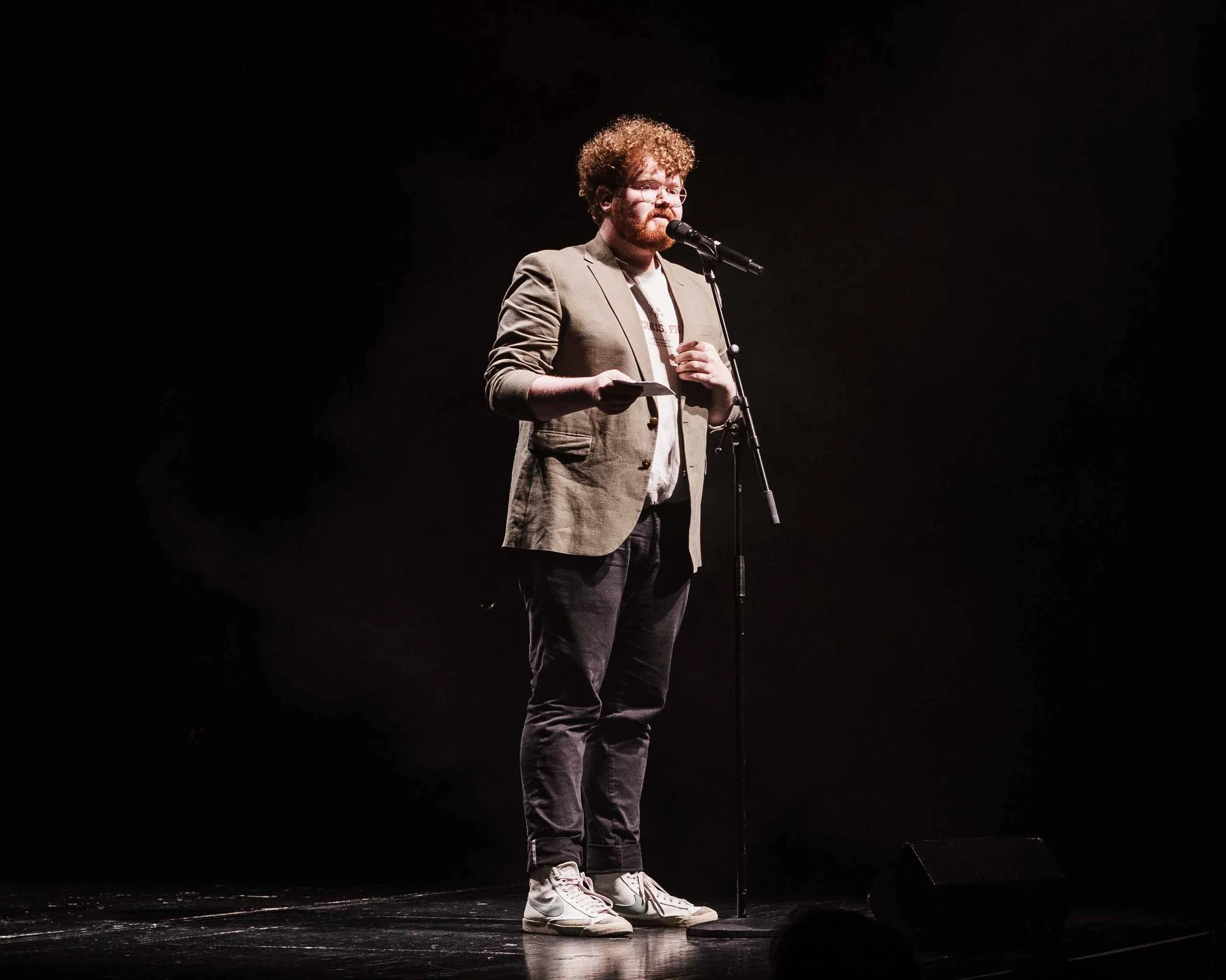 Kolja Fach auf der Bühne spricht in ein Mikrofon, trägt Brille, grau Anzug, weiße Sneakers, dunkelrotes Lockengehaar. Der Hintergrund ist schwarz.