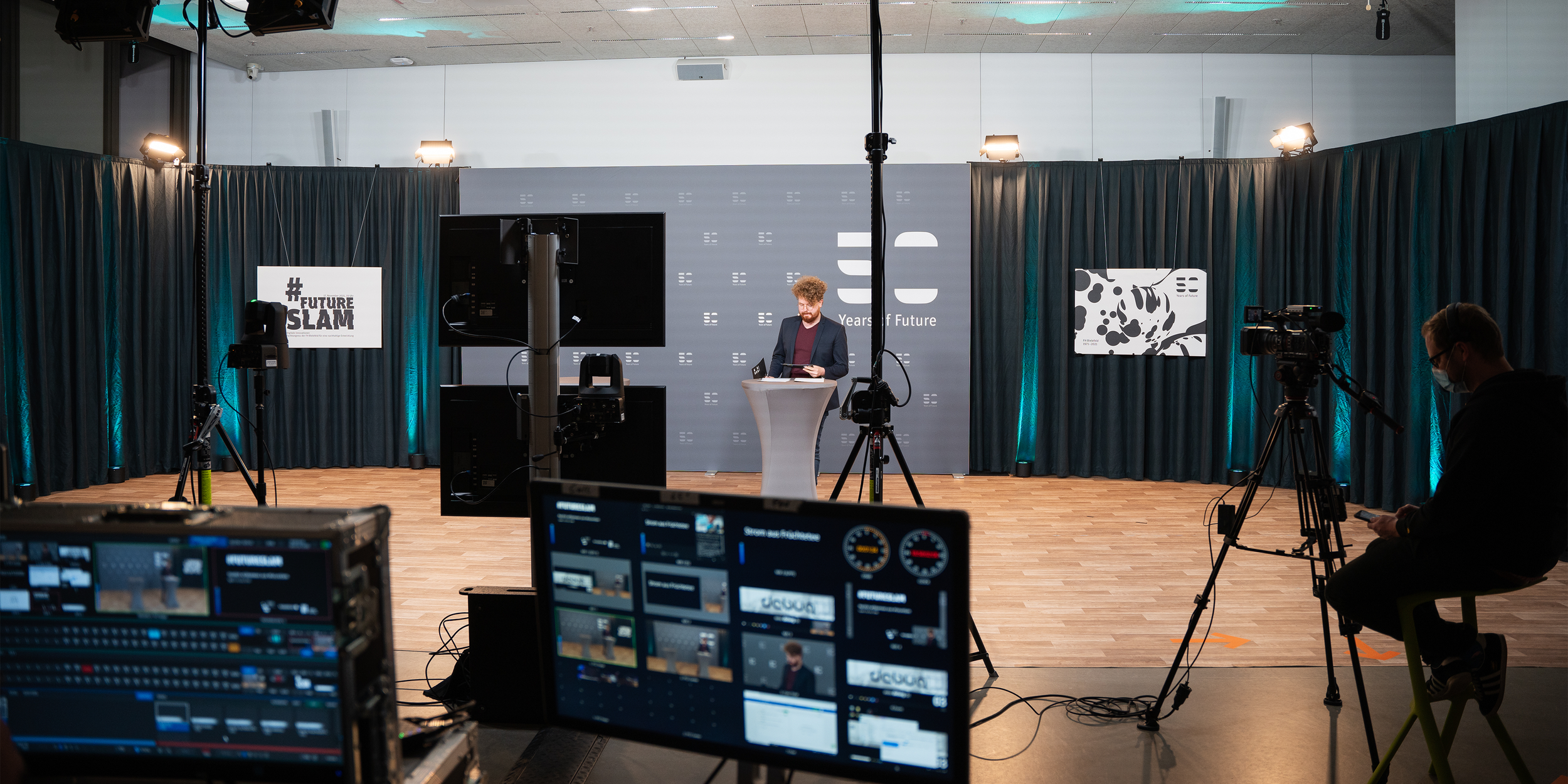 Kolja Fach steht an einem Stehtisch vor einer grauen Bühne und moderiert eine Veranstaltung. Im Vordergrund sind Kameras, Monitore und technische Geräte sichtbar, die den Livestream des Events festhalten.