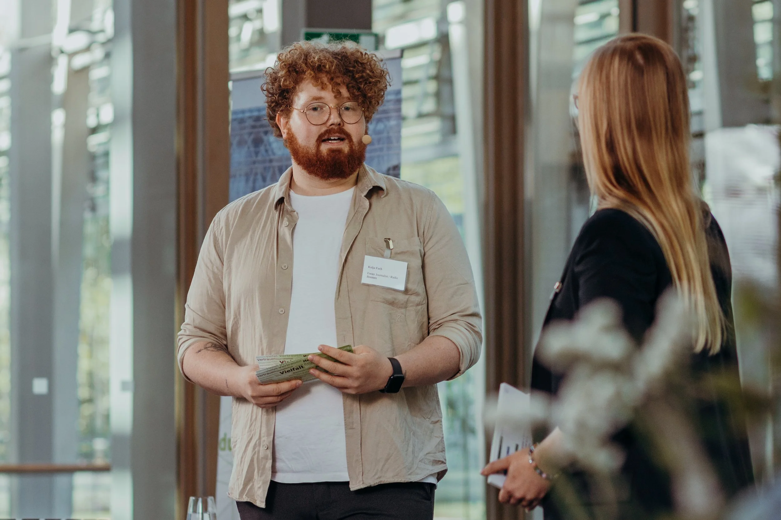 Kolja Fach spricht als Moderator bei einer Veranstaltung. Er trägt eine beige Jacke und hält Moderationskarten in der Hand.