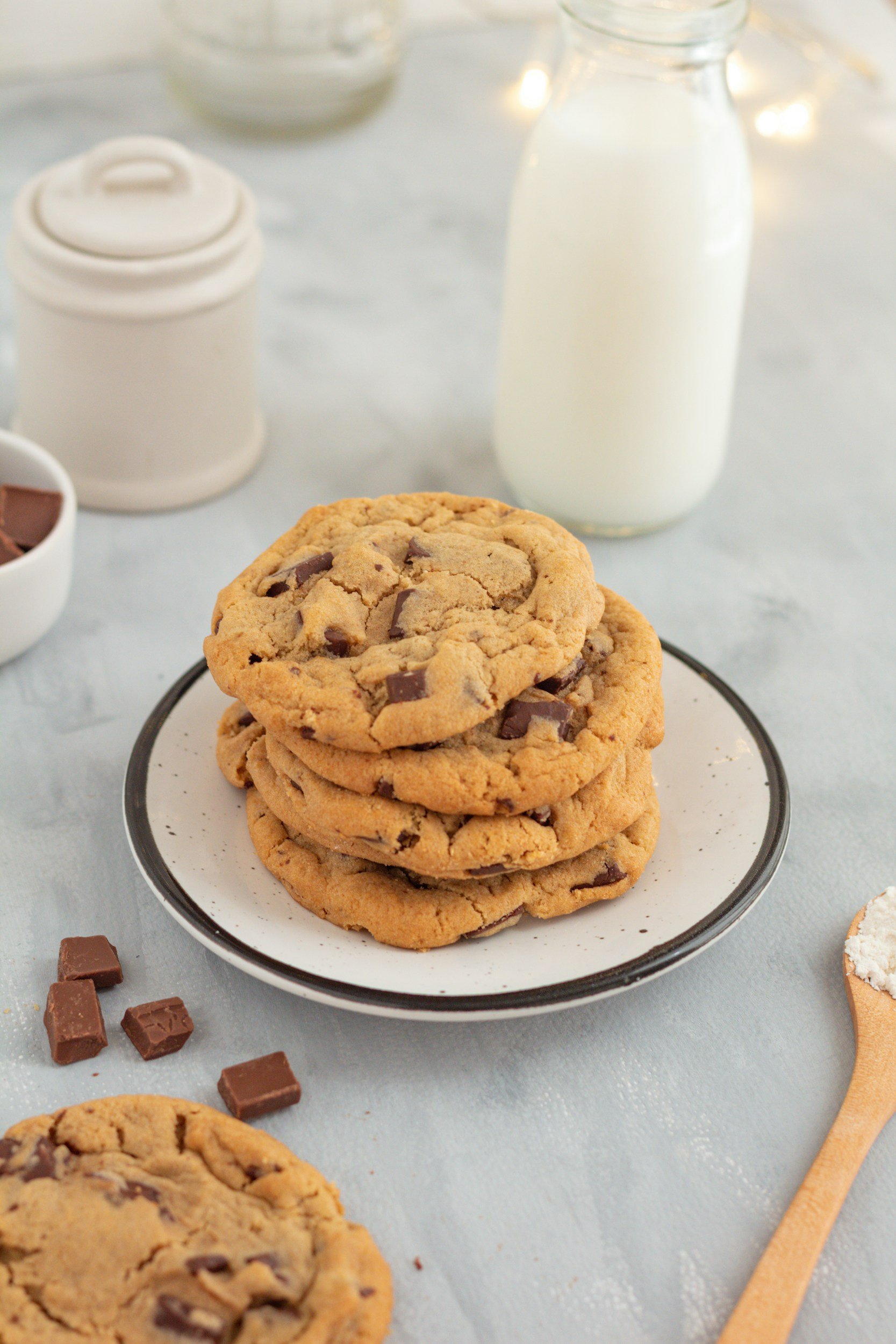 A plate of four chocolate chip cookies on a white dish, with a jar of milk and a small container of chocolate pieces in the background, and a wooden spoon with flour on the right side.