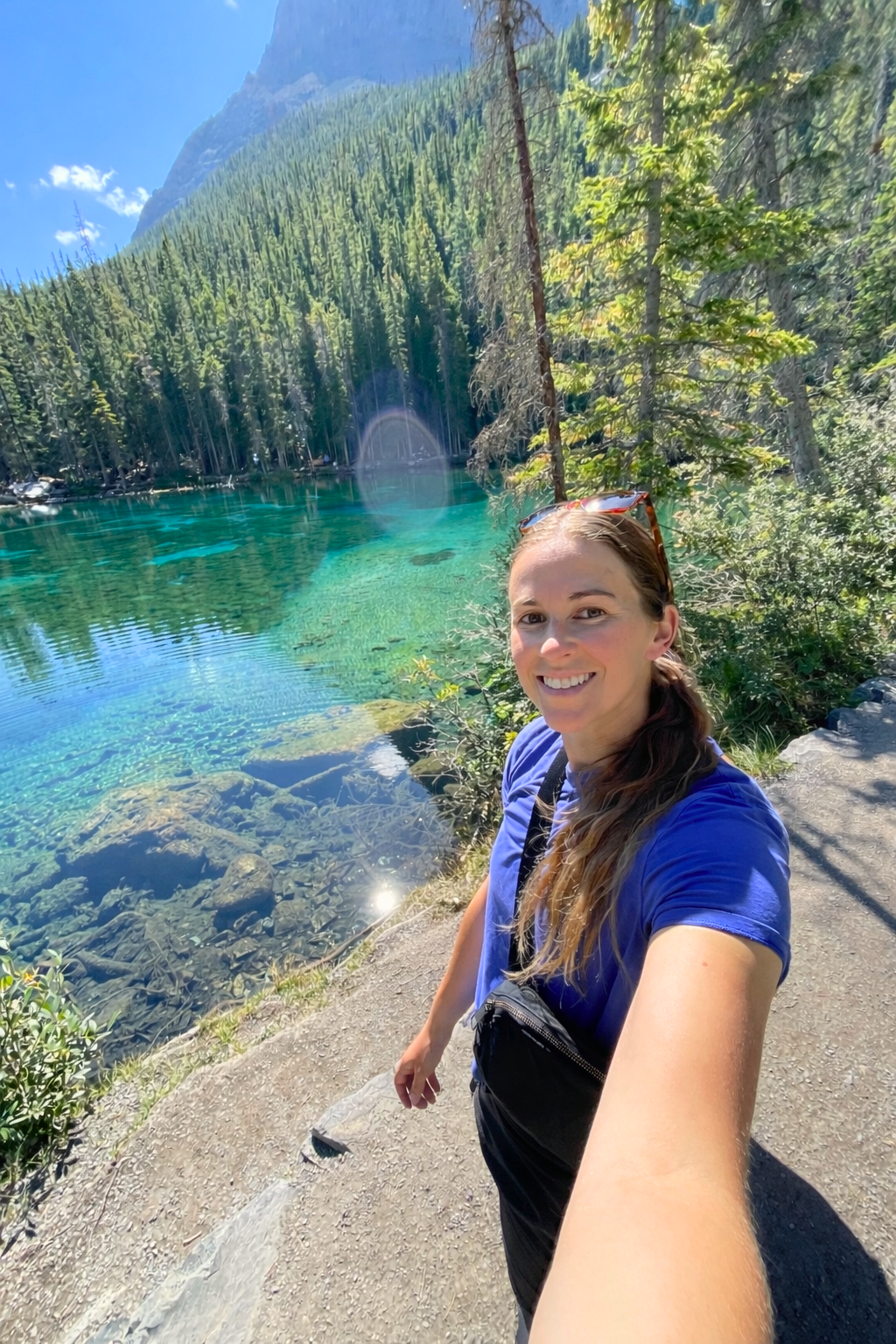 A woman taking a selfie outdoors at a clear, turquoise lake surrounded by green trees and mountains, with a bright blue sky.