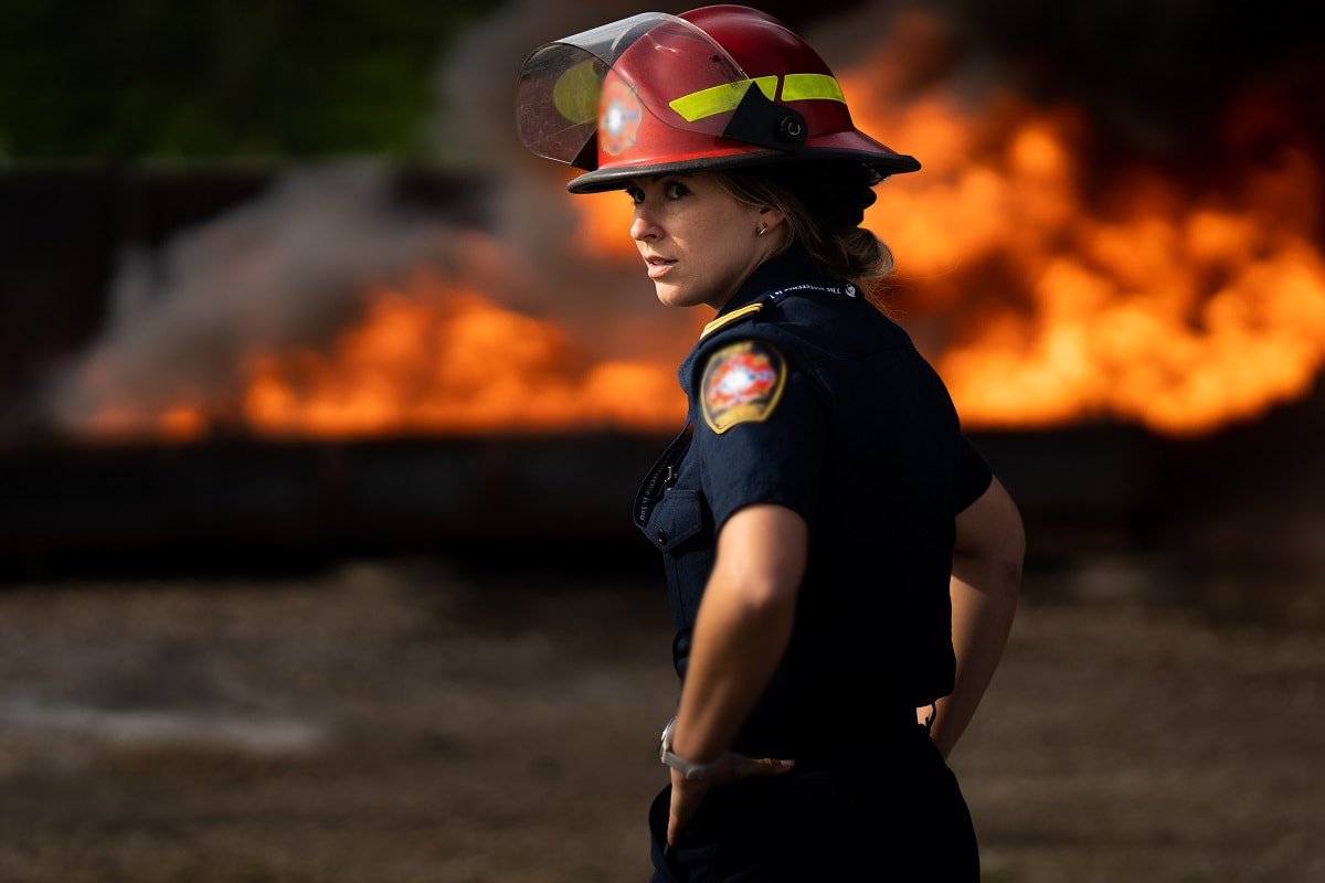 Firefighter in uniform and helmet standing in front of a large fire with flames and smoke.