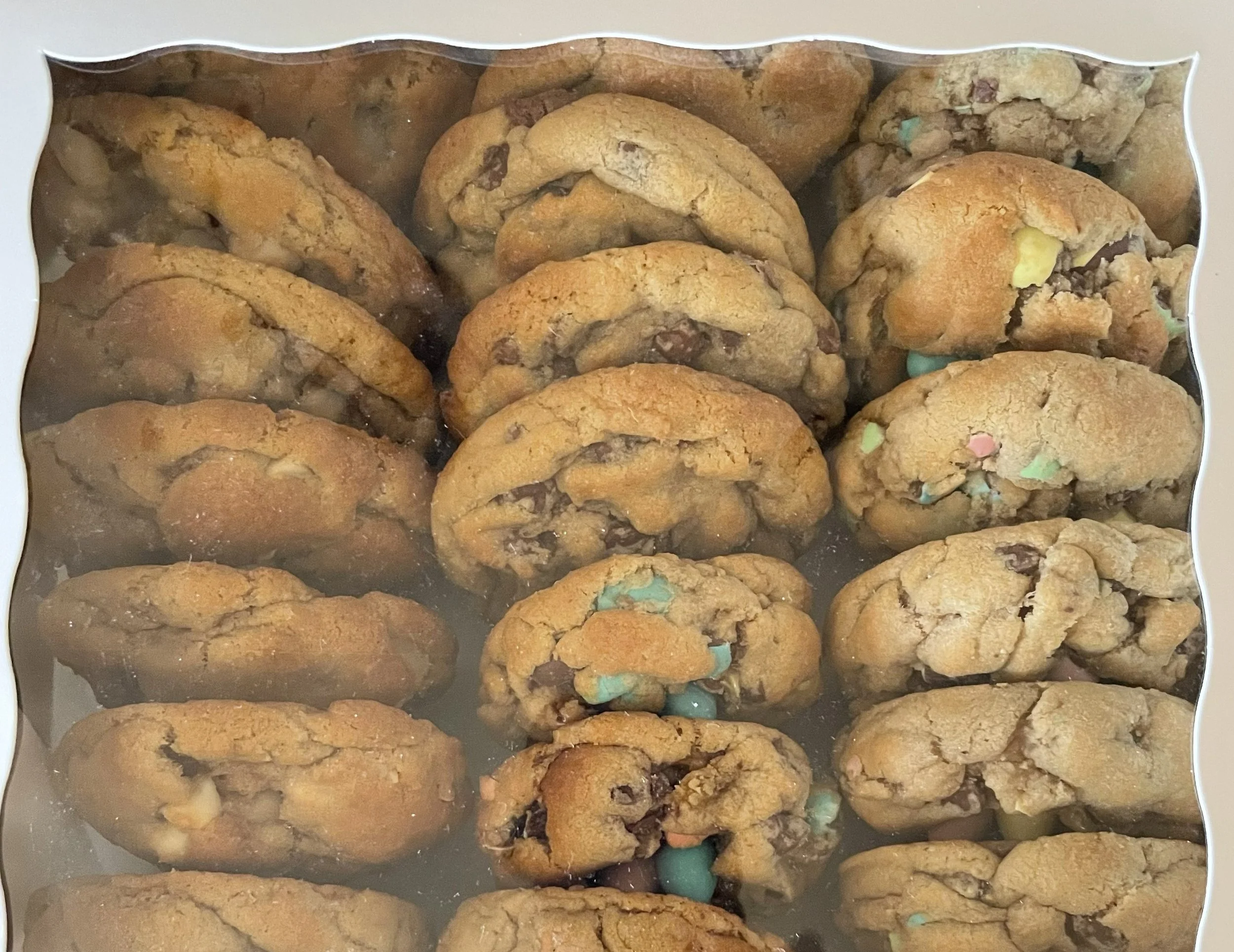 A box of assorted chocolate chip cookies with colorful candies inside, viewed through a clear plastic window.