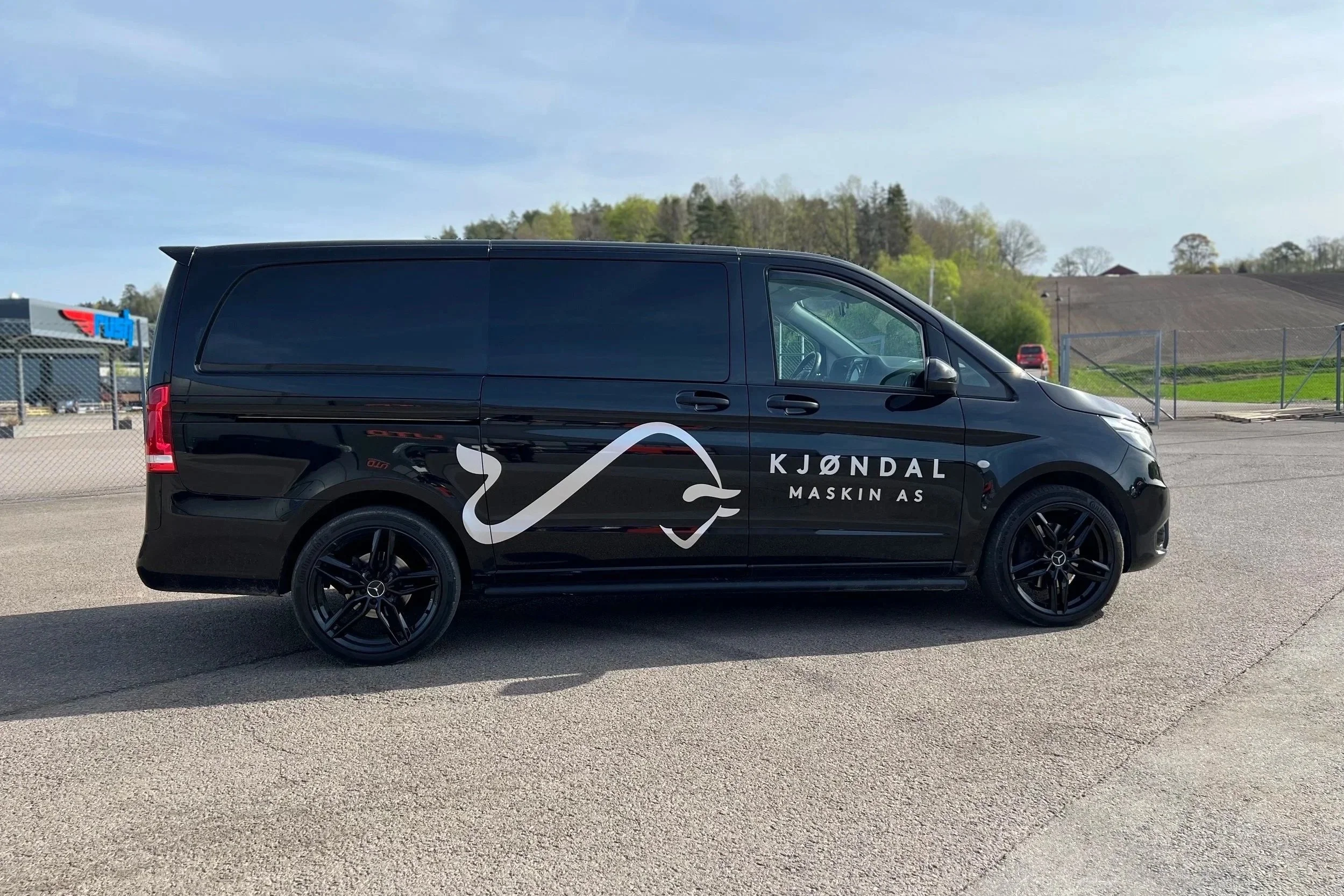 Svart varebil med logo og teksten "KJØNDAL MASKIN AS" på siden, parkert på asfalt med en bakke og trær i bakgrunnen.