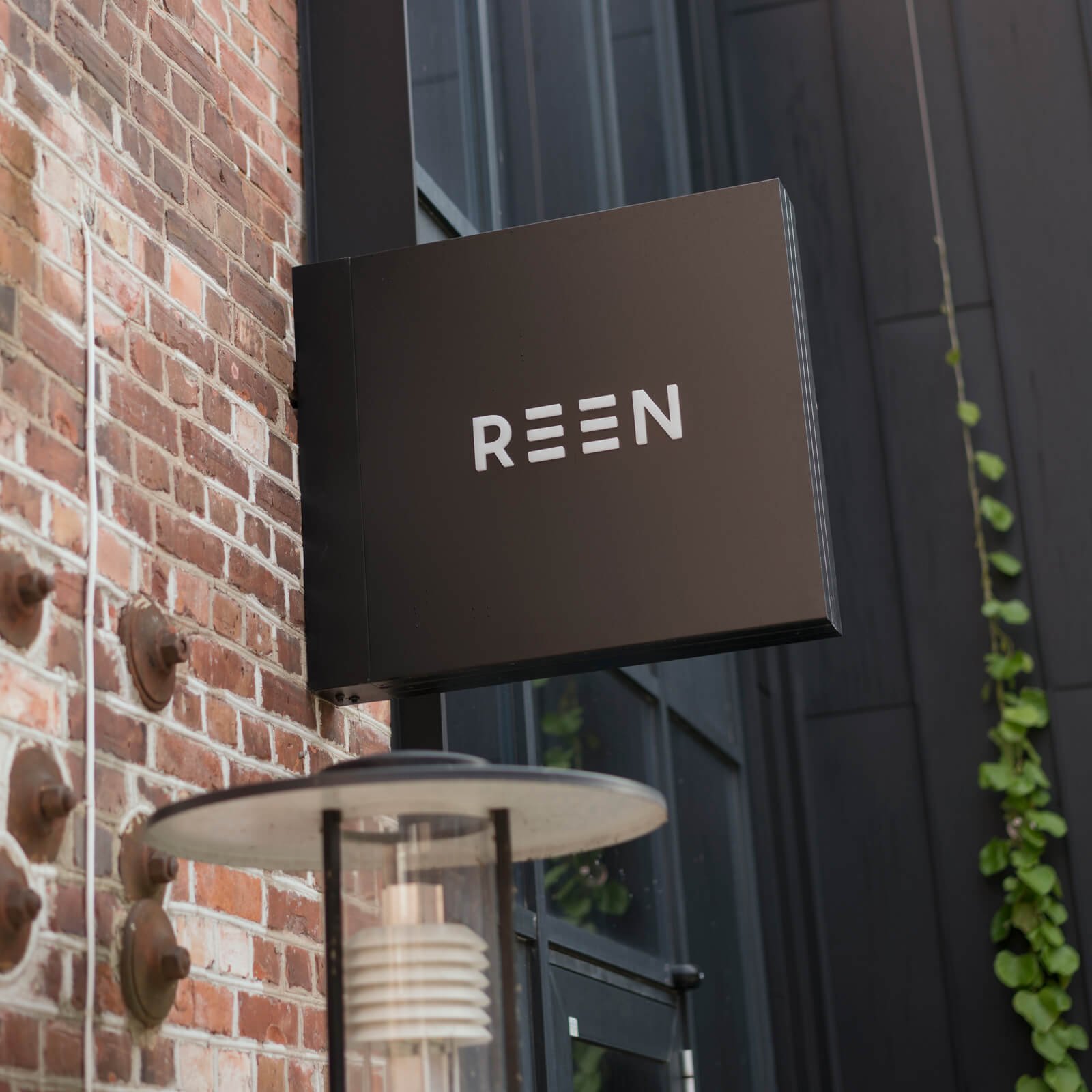 Et skilt med teksten 'REEN' montert på en vegg utenfor en bygning, omgitt av en murvegg og noen grønne planter.