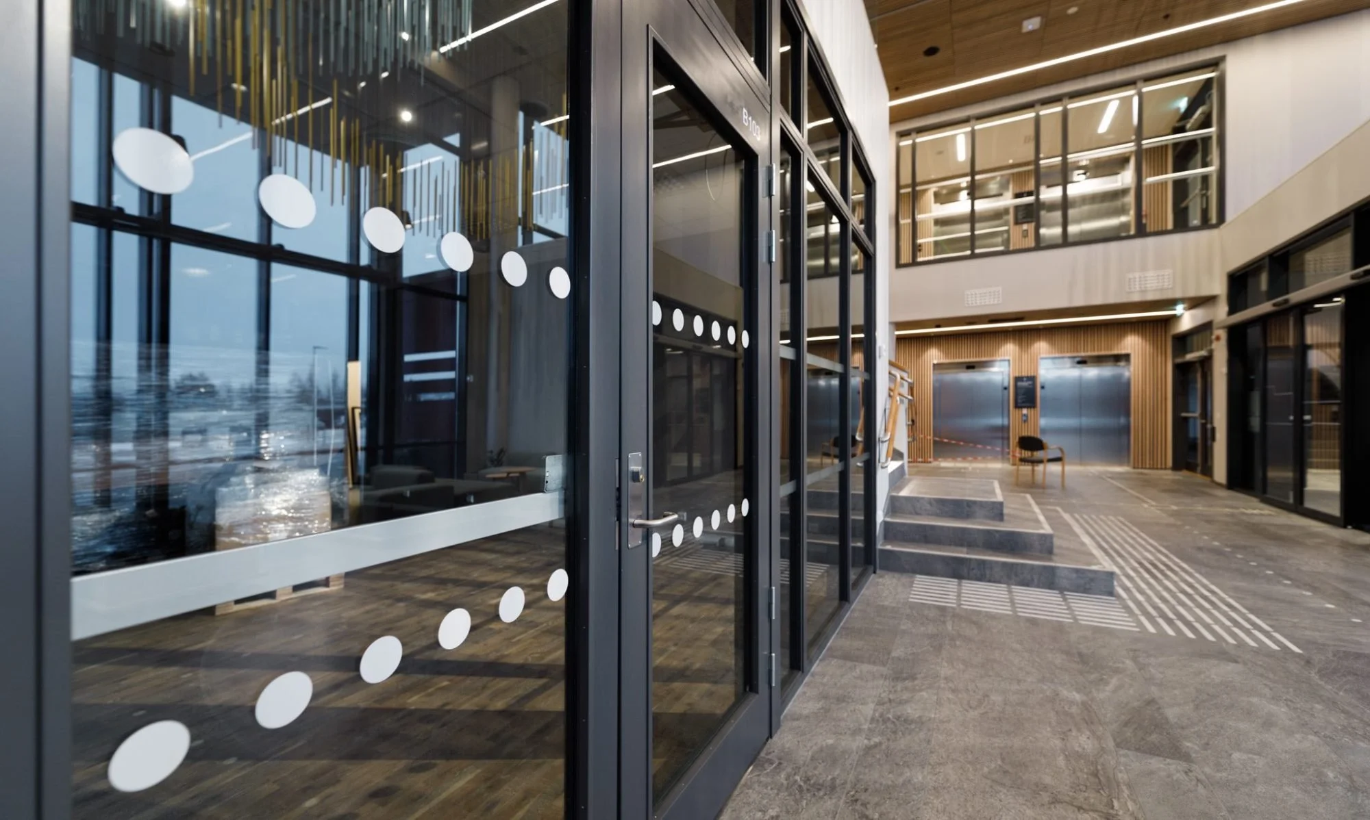 Innvendig entré i en moderne bygning med glassdører, stoler og heis i bakgrunnen, trevegger og store vinduer.