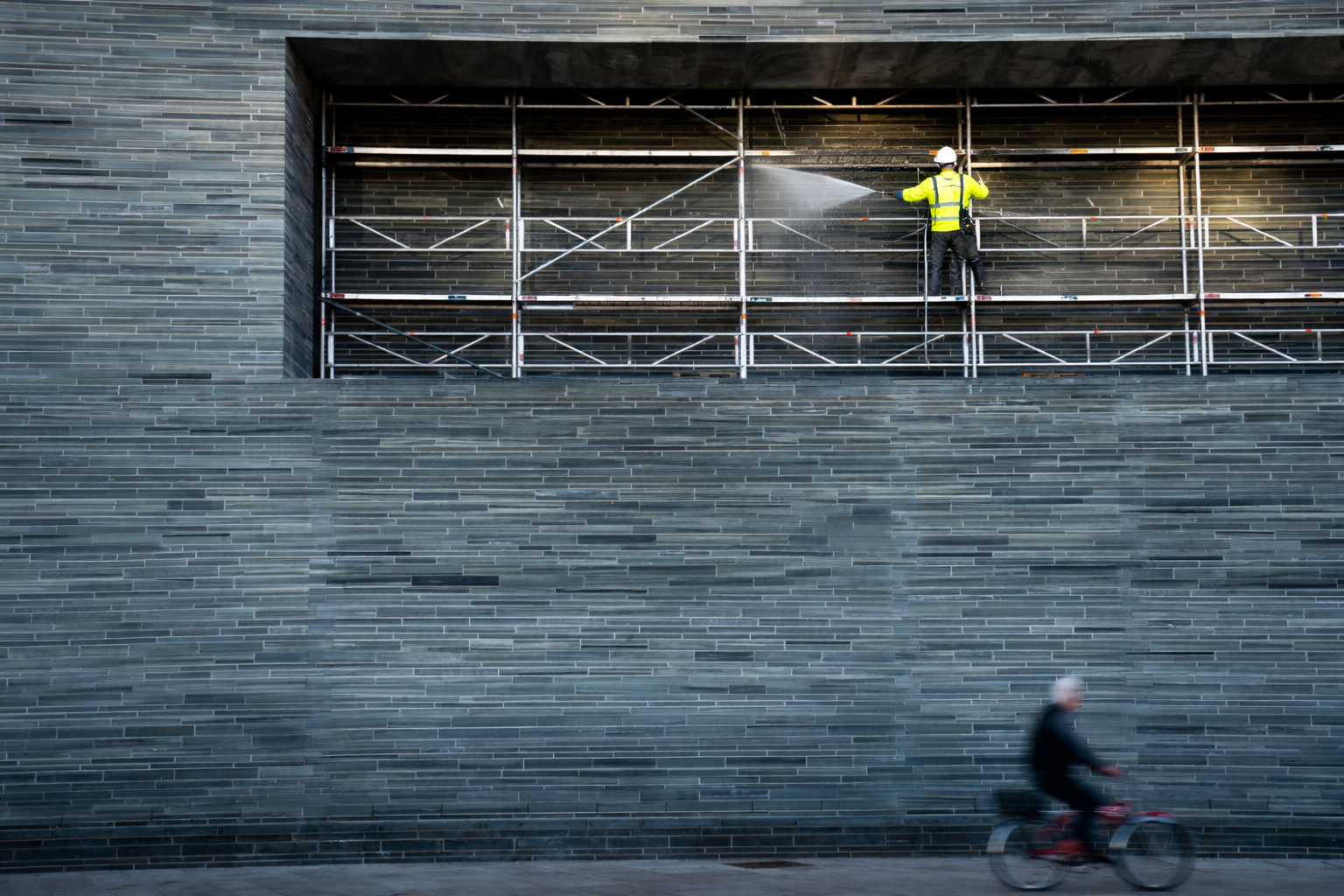 En mann i gul vest og hjelm vasker en vegg på en byggeplass, mens en person sykler forbi.