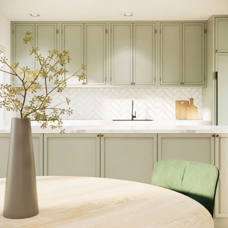 Et lyst kjøkken med grønne skap, hvit benkeplate og to skjærebrett av tre. I forgrunnen er det en rund bordflate med en grå vase med kvitt blader.