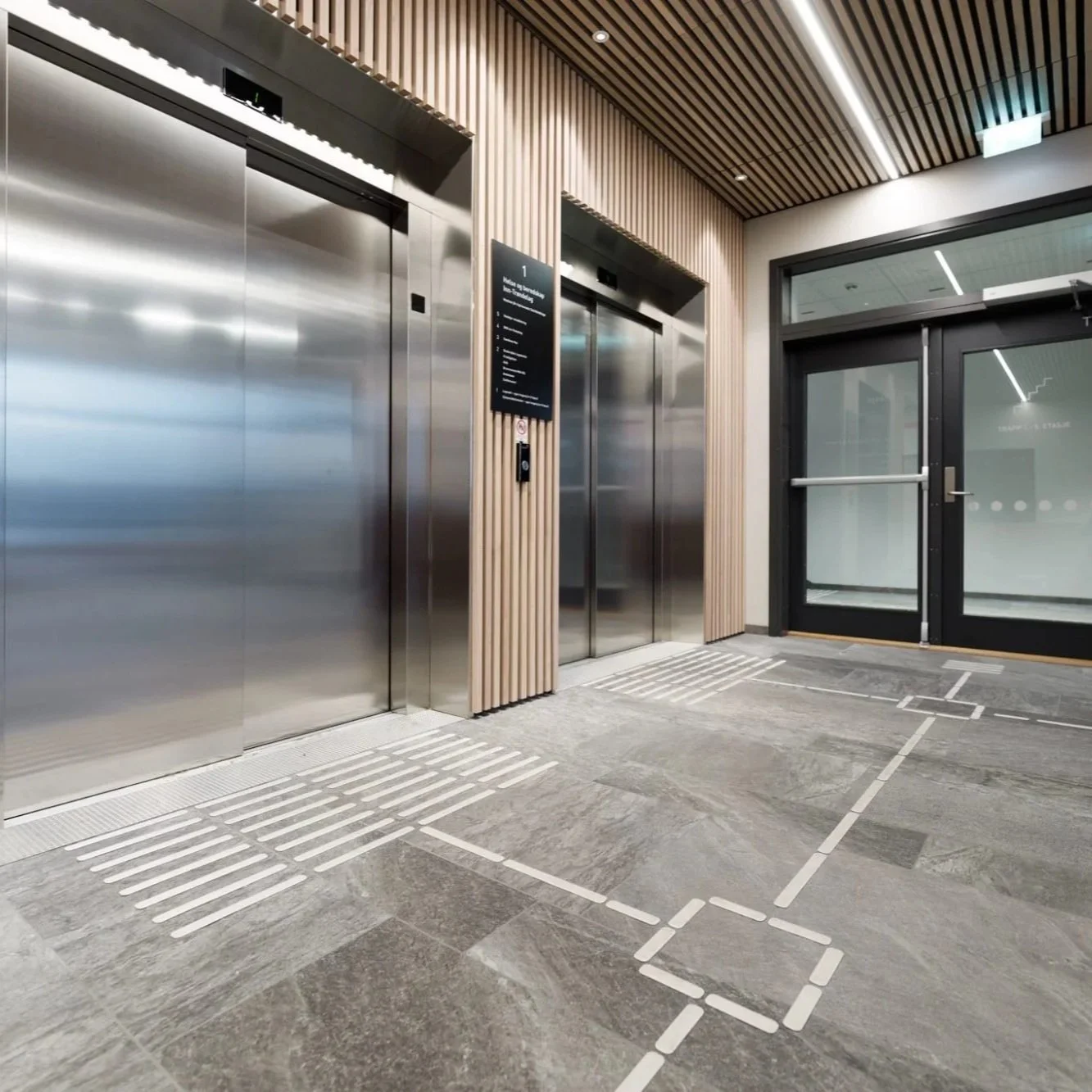 Innendørs bilde av to heiser med metallskjolder og en glassdør til høyre. Golvet har markeringer for tilgjengelighet.