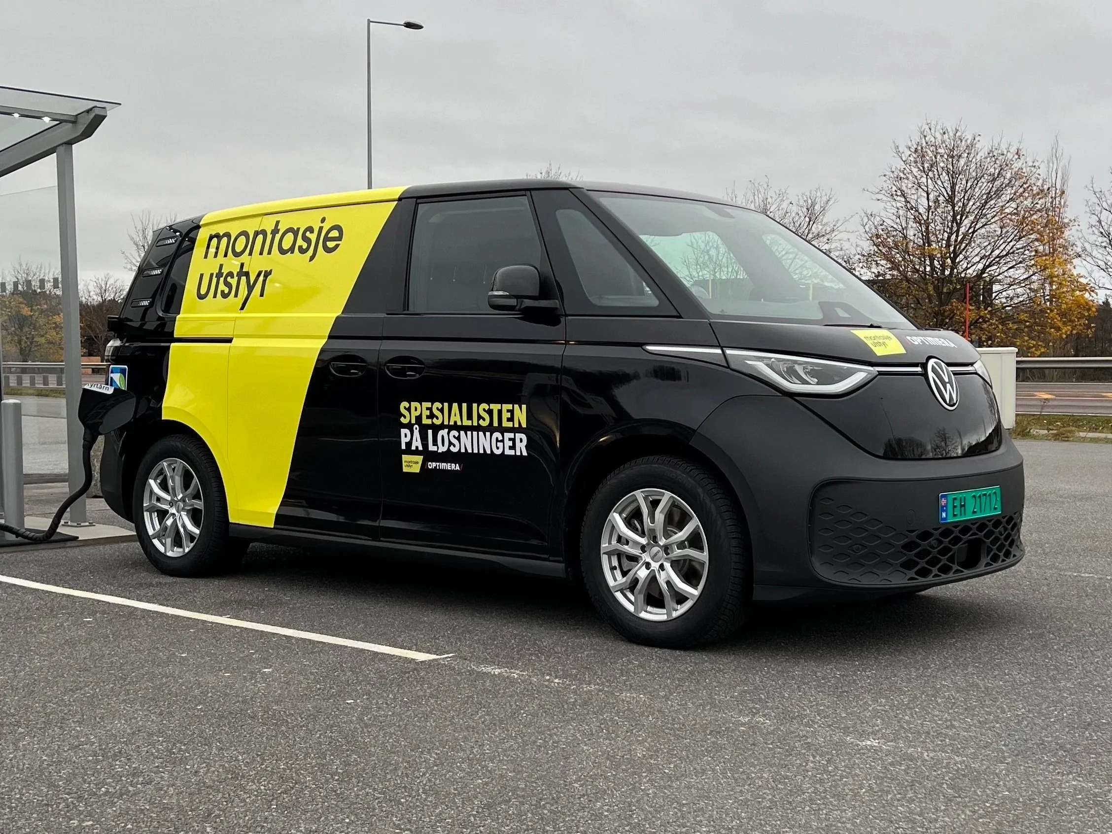 En svart Volkswagen elektrisk varebil som lades på en ladestasjon. Bilen har gul og svart reklame med teksten 'montasje utstyr' og 'SPESIALISTEN PÅ LØSNINGER'.