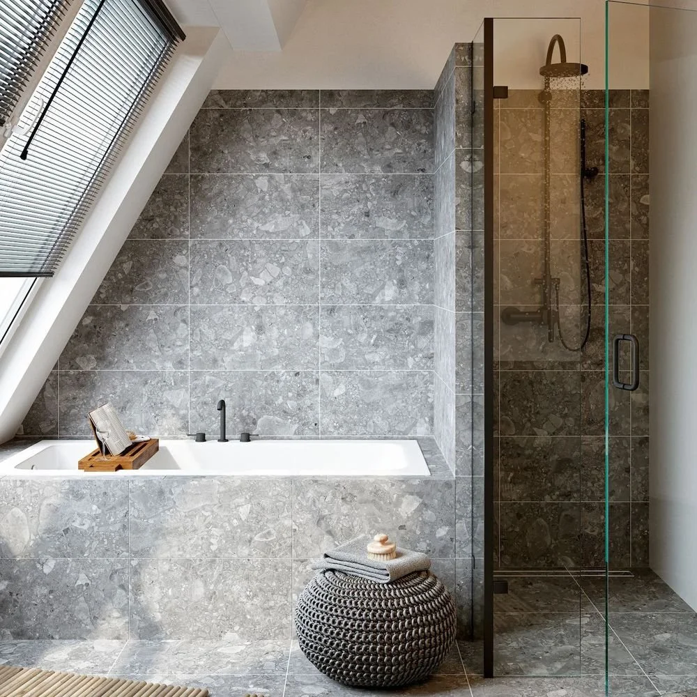 Moderne bad med badekar, vegg og gulv dekket med grå steinfliser, stor vindu med persienner, og en separat dusj med glassvegg.