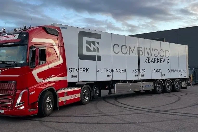 En stor, rød lastebil med en lang, hvit trailer med logo og tekst for Combiwood Barkevik. Trailerens side viser flere ord på norsk, inkludert 'istverk', 'utforminger', 'spiler', 'panel', og nettsiden combiwood.no.
