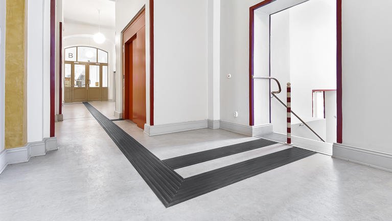 Interiør av et trappeområde i en bygning med hvite vegger, svart teppetrand rundt trappen, og en utgang til trapperom.