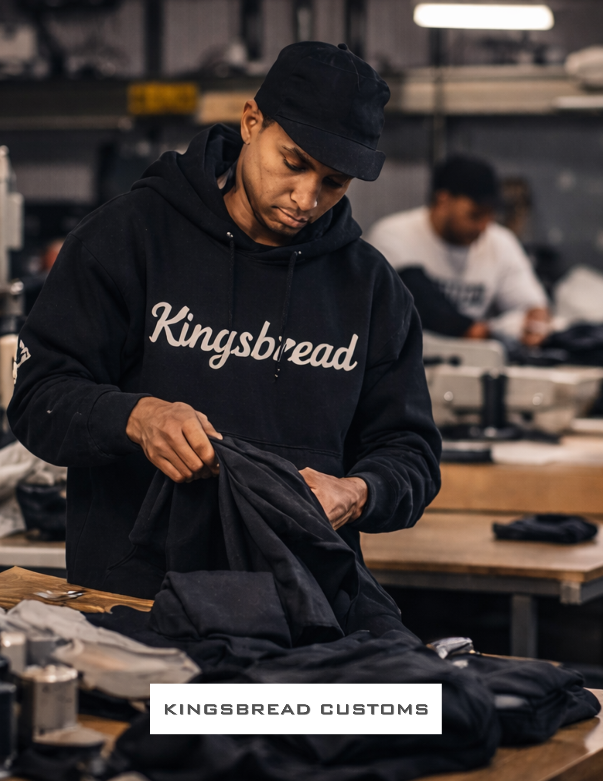 A man wearing a black hoodie with 'Kingsbread' printed on it, working on clothing at a workshop table with another person in the background.