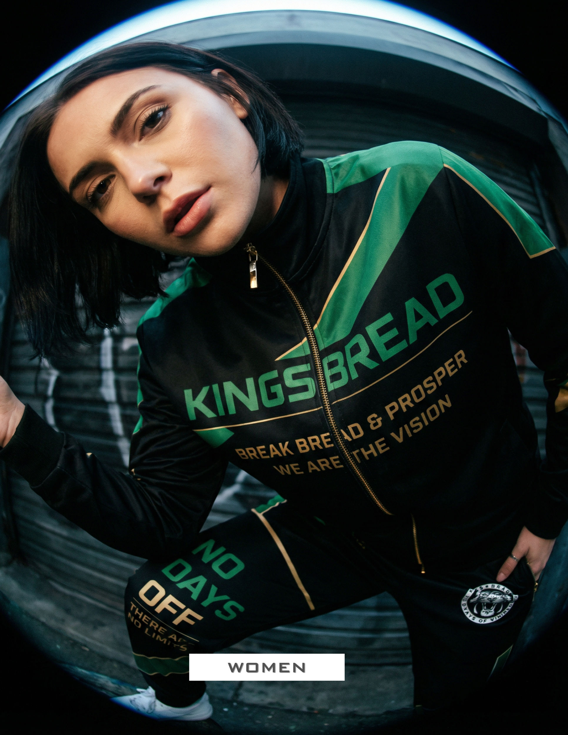 A woman with short dark hair wearing a black sports jacket with green and gold accents, with the words 'Kings Bread' and 'Break Bread & Prosper' printed on it, leaning forward and looking at the camera.
