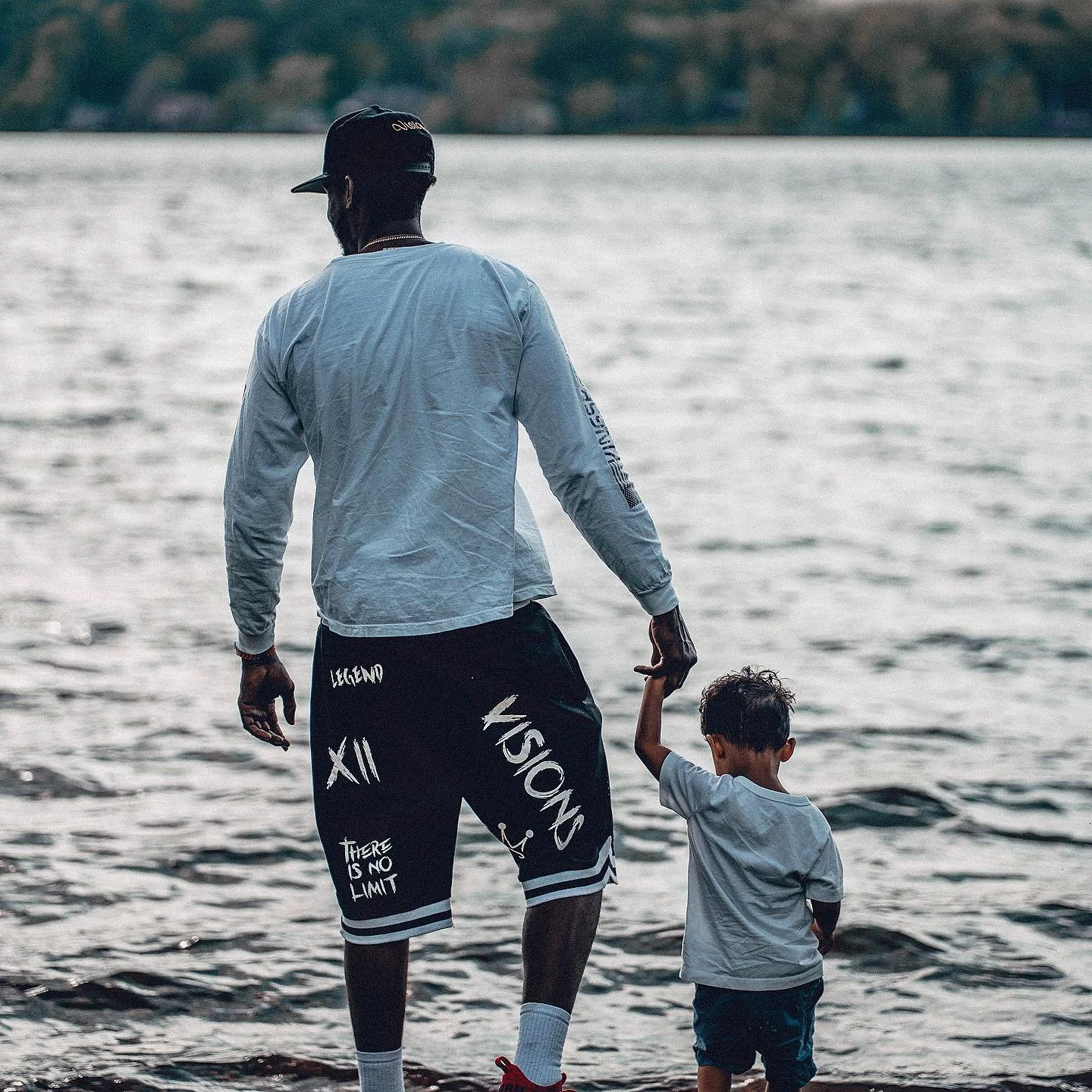 An adult man and a young boy walk hand in hand along the edge of a body of water at sunset or dusk.