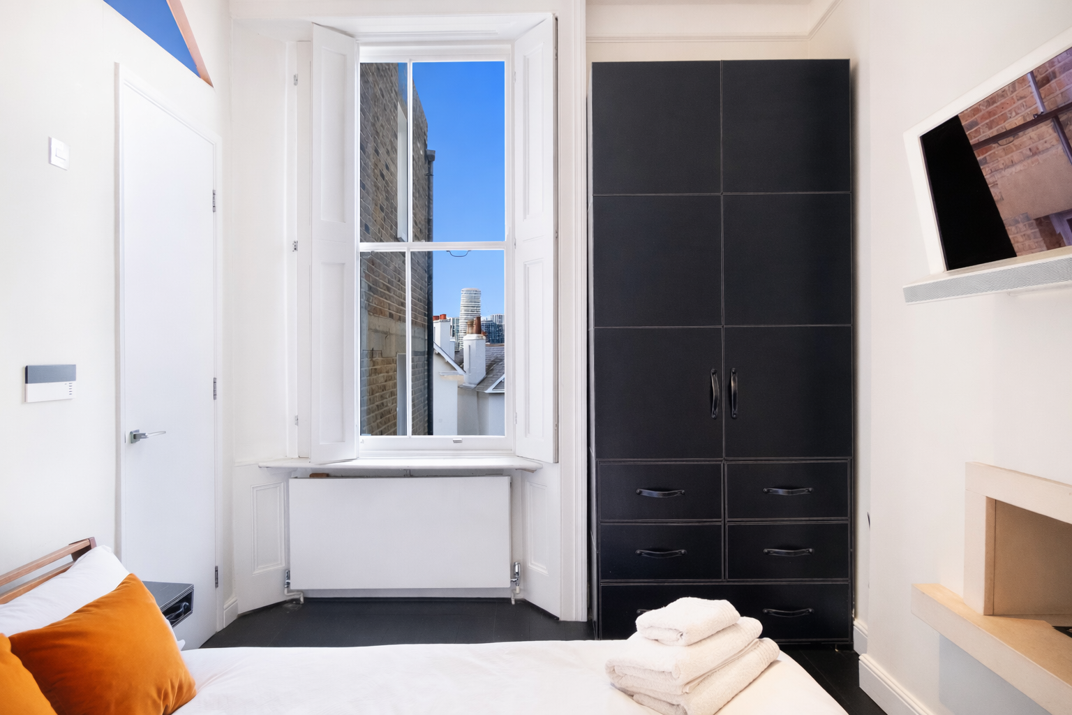 Bright modern bedroom with open window, black built-in wardrobe, white bed with orange pillow, folded towels, and wall-mounted TV