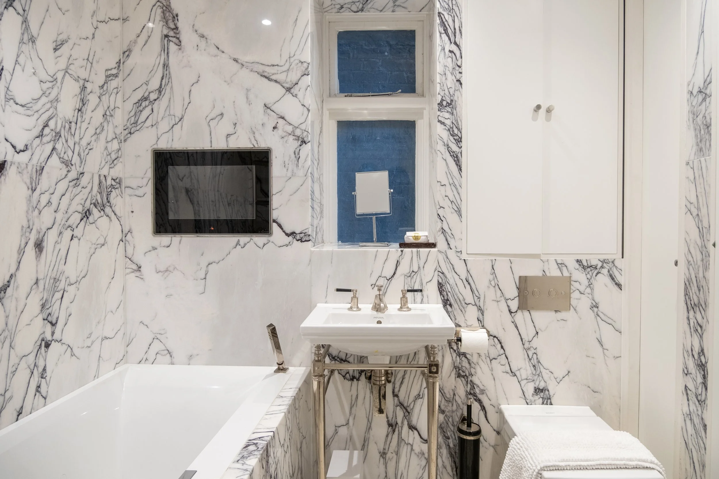 Elegant marble bathroom with freestanding bathtub, pedestal sink, wall-mounted television, window, and toilet