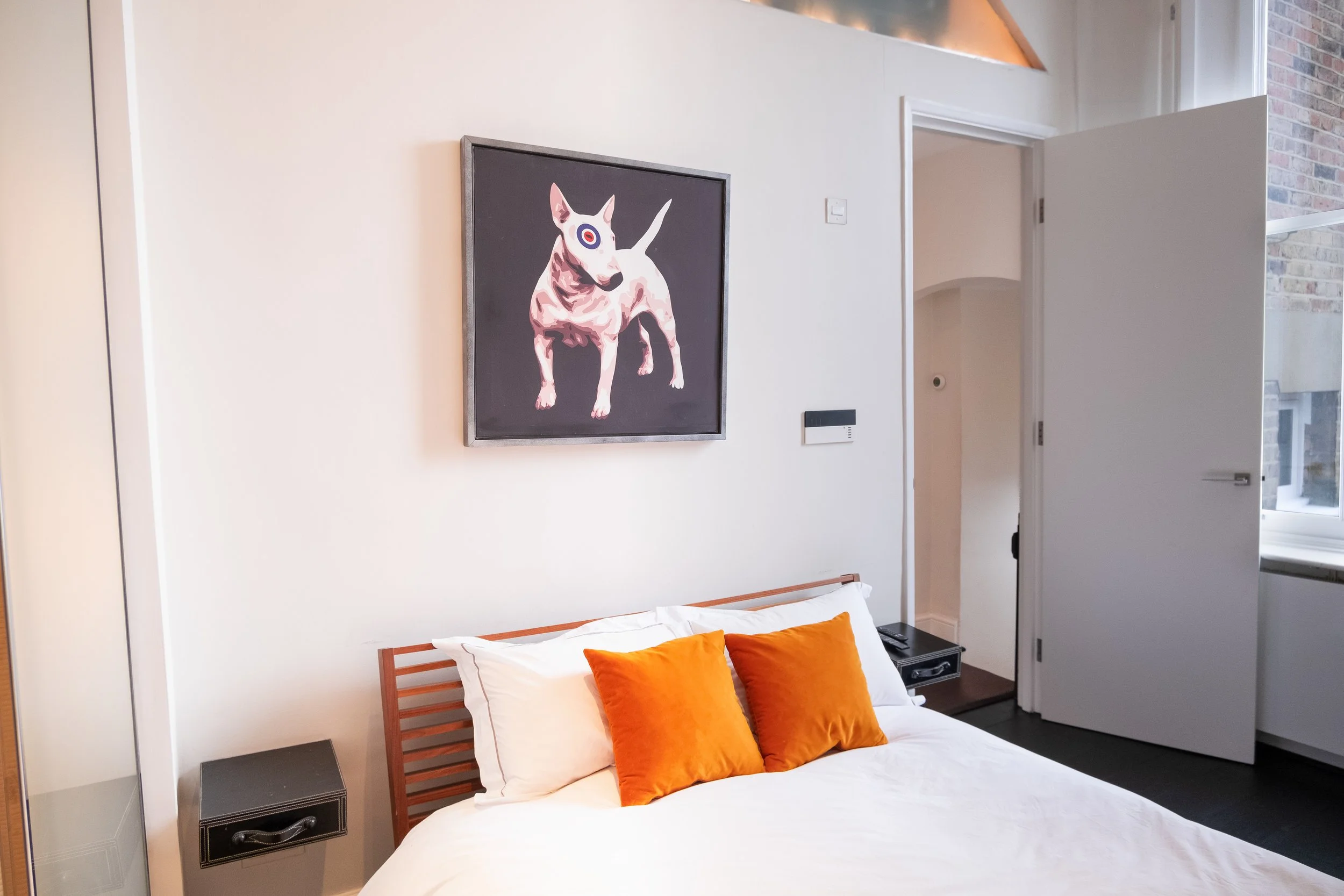 Modern bedroom with white bedding, orange accent pillows, black nightstands, and framed abstract dog artwork