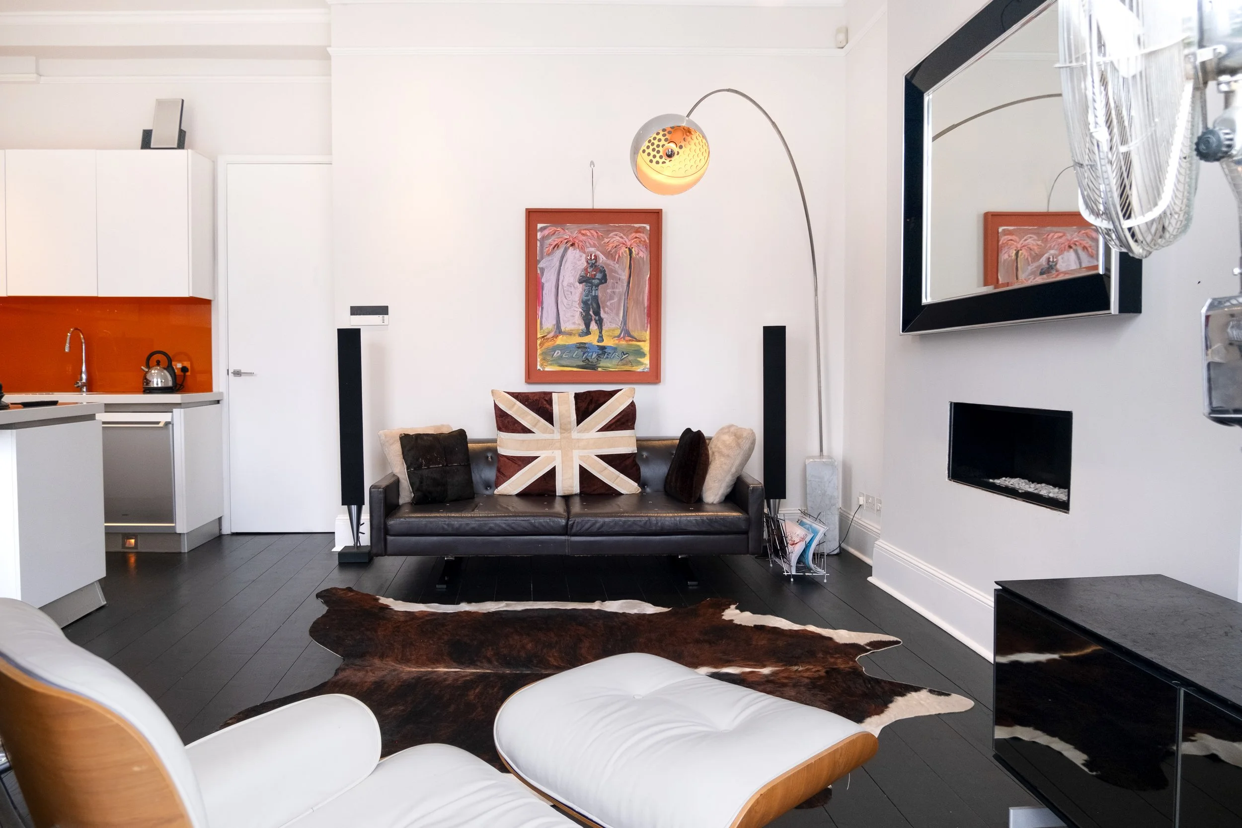 Modern living room with black leather sofa, Union Jack cushion, cowhide rug, wall-mounted TV, fireplace, floor lamp, and adjoining kitchen.