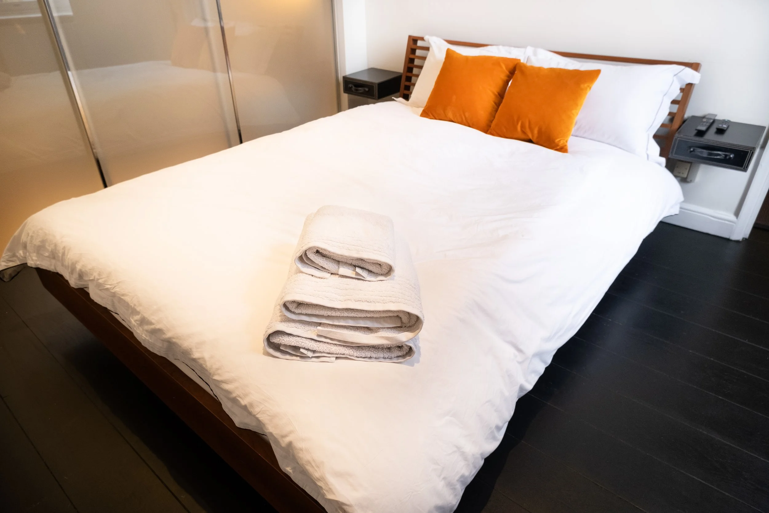 Modern bedroom with a white bed, orange accent pillows, folded towels, bedside tables, and mirrored closet doors