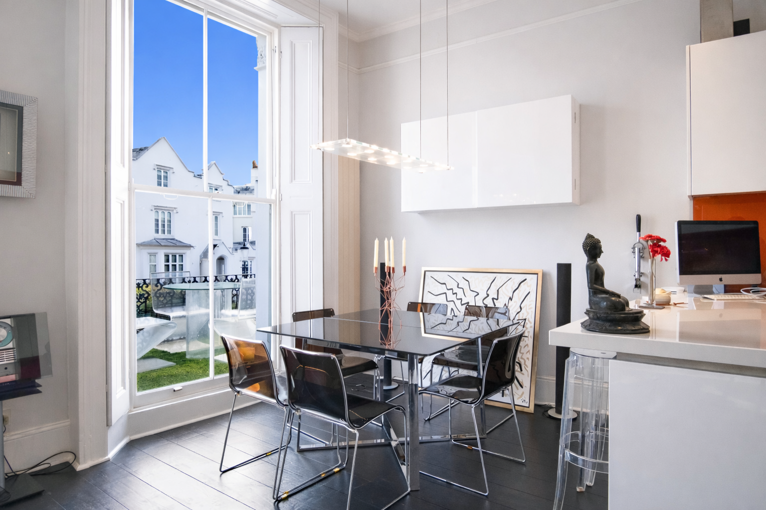 Modern white dining area with glass table, black chairs, pendant light, large window, artwork, and kitchen counter