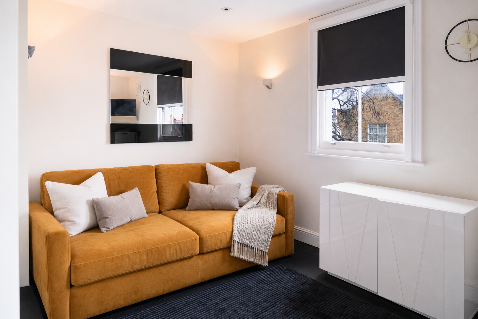Modern living room with a mustard-yellow sofa, white pillows, throw blanket, black rug, wall mirror, and white cabinet beneath a window.