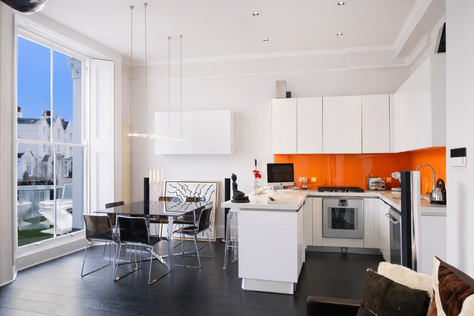 Modern white kitchen and dining area with orange backsplash, central island, black glass table, and large bay window
