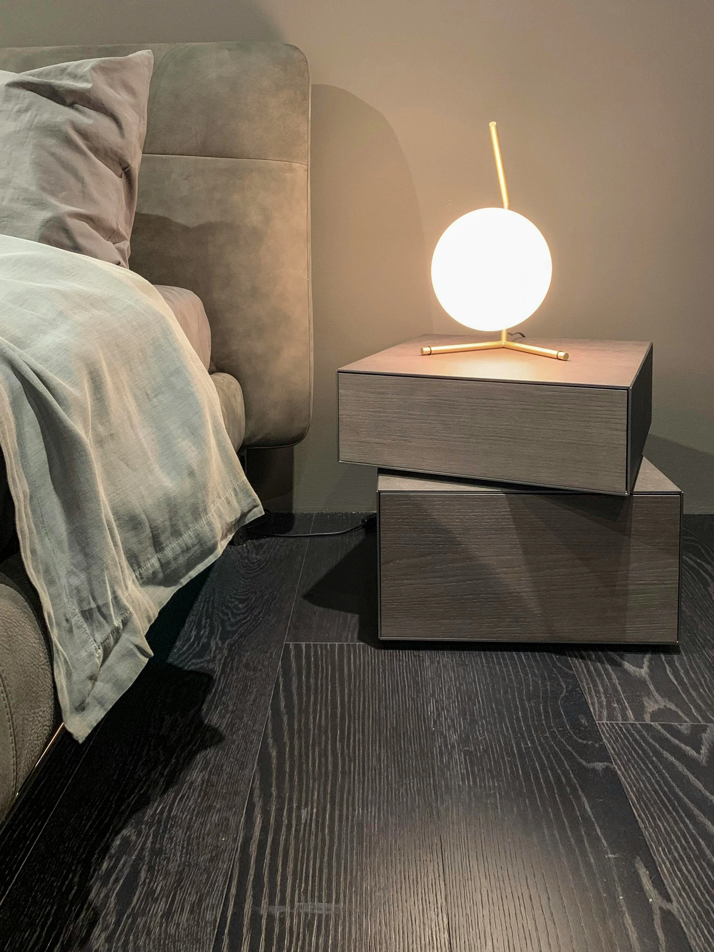 Modern bedroom nightstand with a glowing spherical lamp and a bed with gray bedding and pillows, against a gray wall and dark wooden floor.