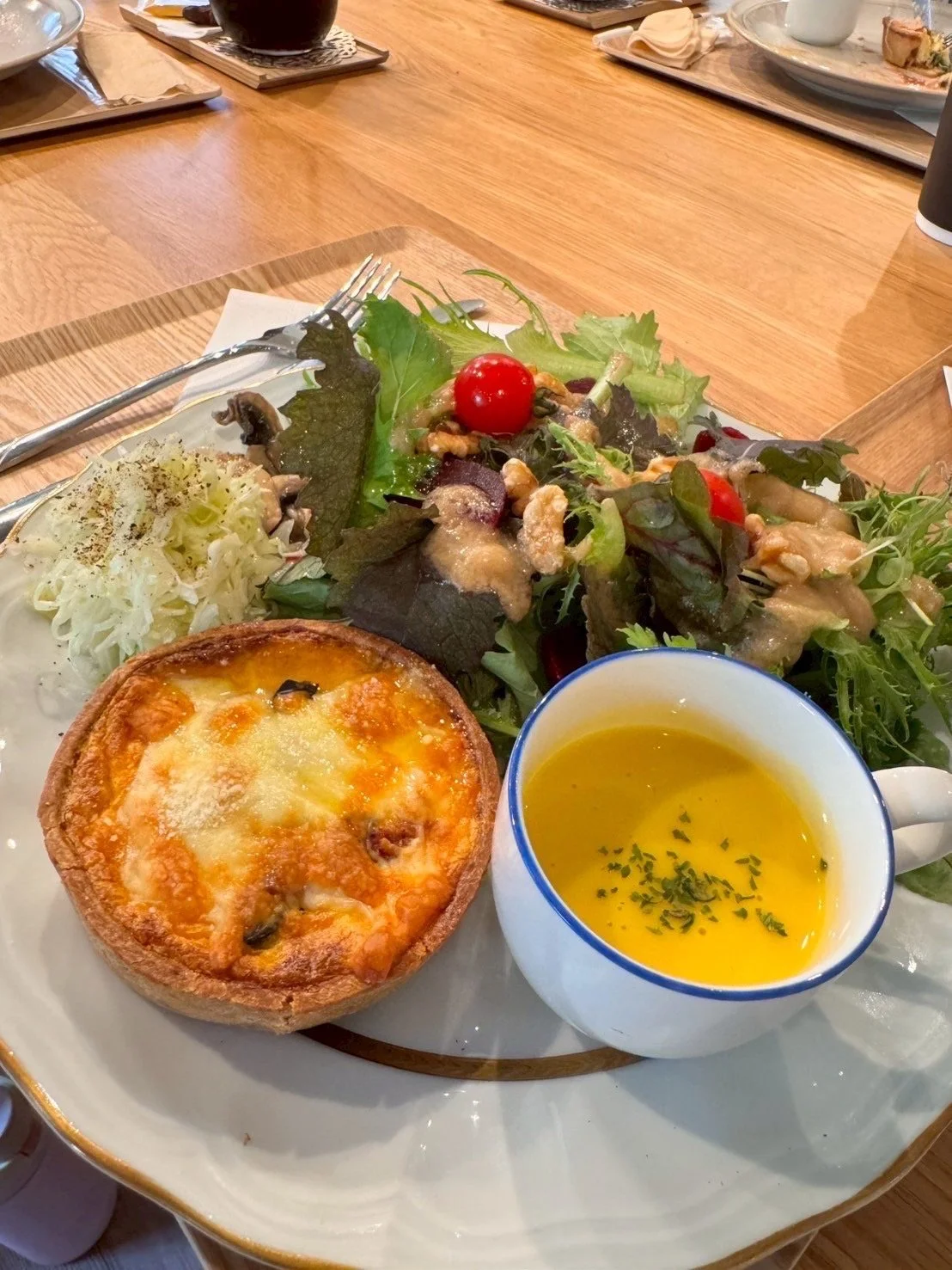 A plate with a small vegetable quiche, a bowl of yellow soup garnished with herbs, and a mixed green salad with cherry tomatoes and nuts on a wooden table.