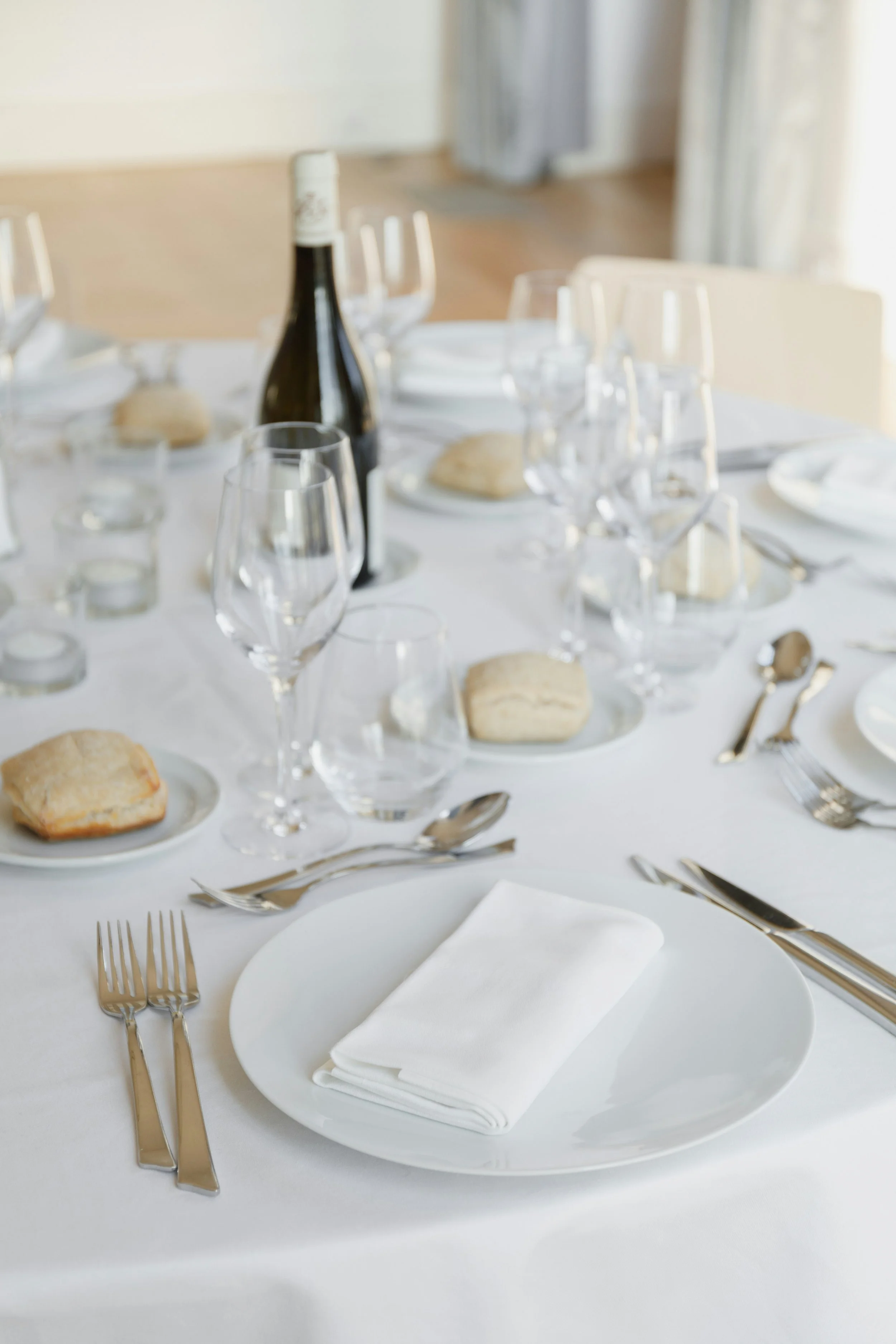 Formal dining table set with white tablecloth, white napkin on a plate, multiple wine glasses, water glasses, cutlery, bread rolls on small plates, and a bottle of wine or champagne in a well-lit room.