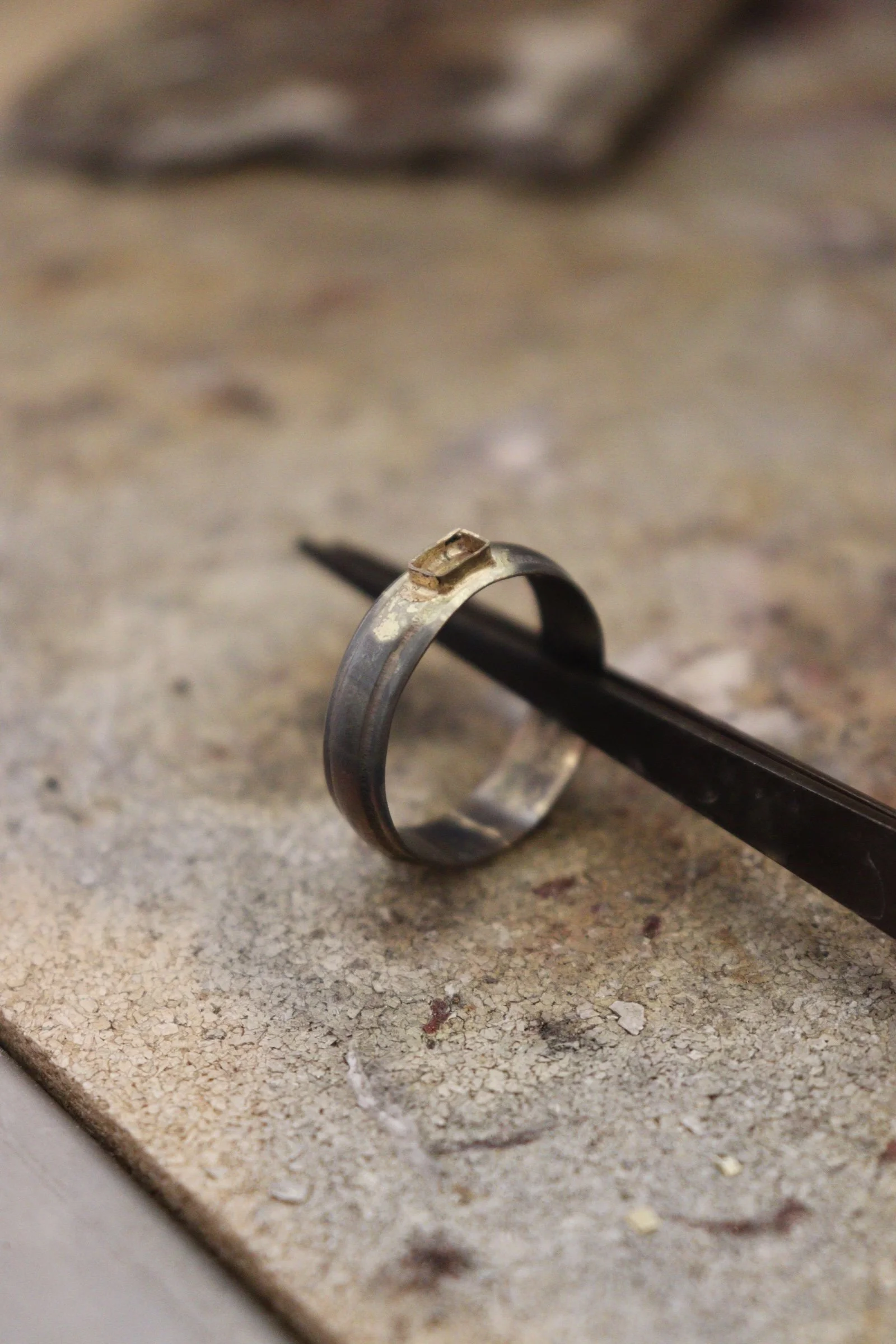 A metal ring with a small decorative element on it, resting on a textured surface with a pair of black tweezers underneath.
