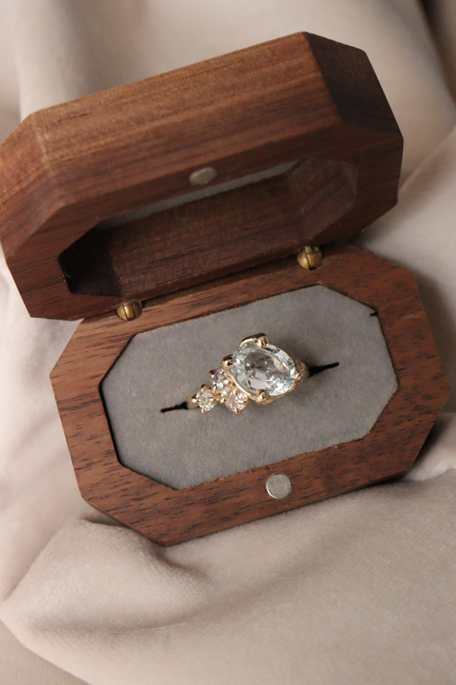 A wooden jewelry box with a hinged lid, containing a ring with a large central clear gemstone and smaller surrounding gemstones, set on a soft fabric surface.