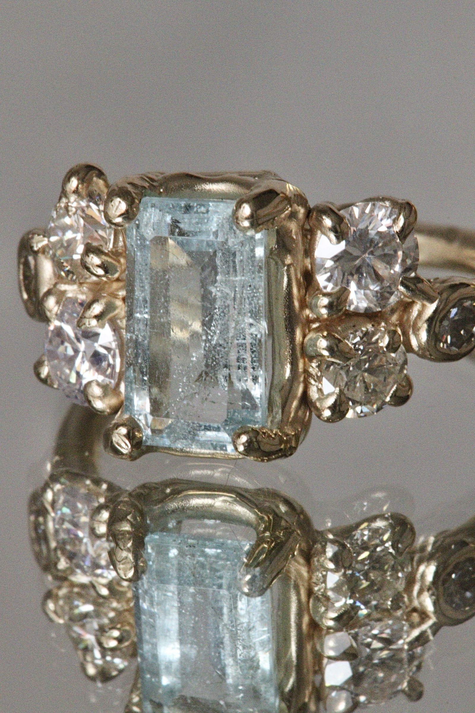 Close-up of a silver ring with a rectangular light blue gemstone and multiple small round diamonds on each side, reflecting on a mirror surface.