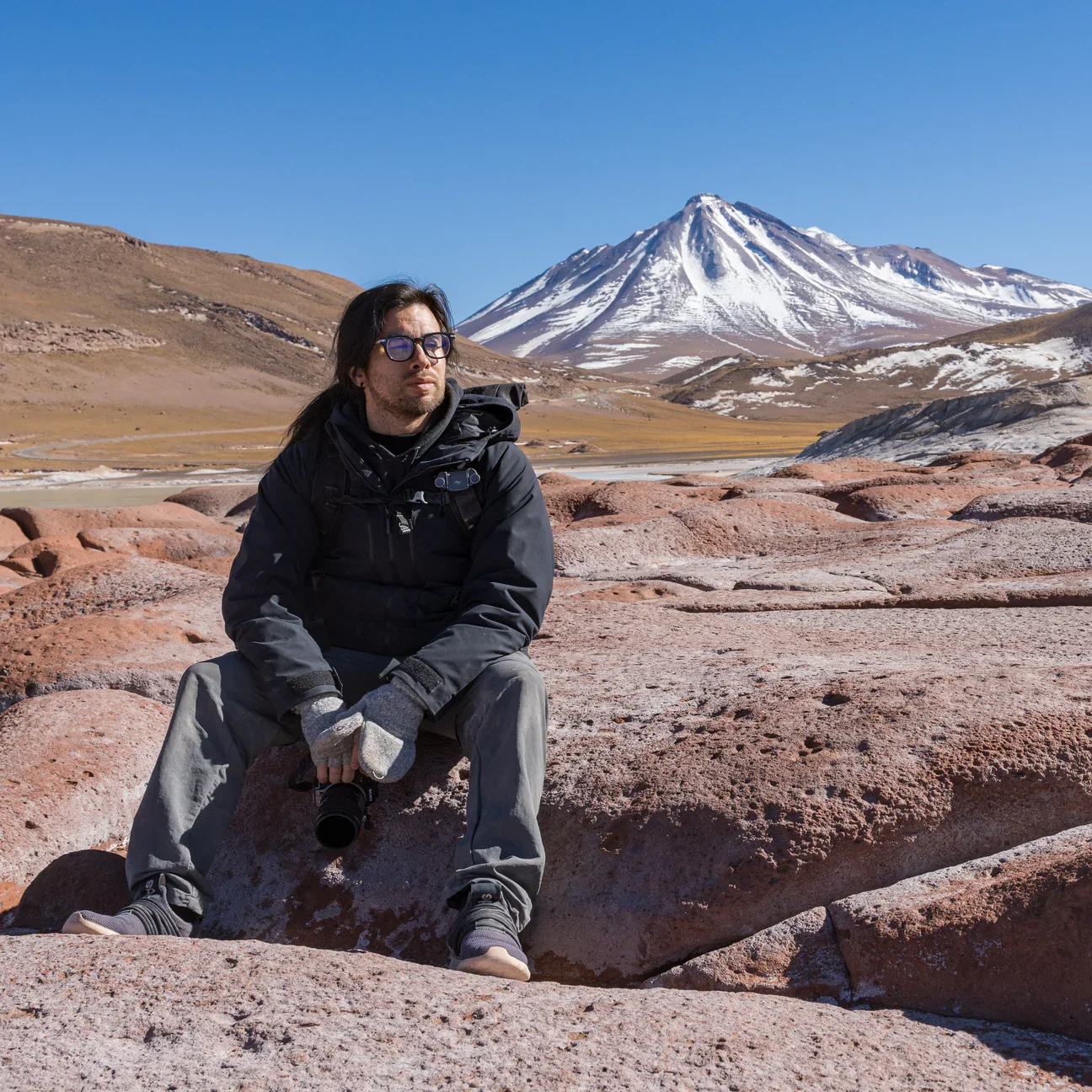 A man sitting on rocks in a mountainous area with snow-capped volcano in the background, wearing outdoor gear and holding a camera.