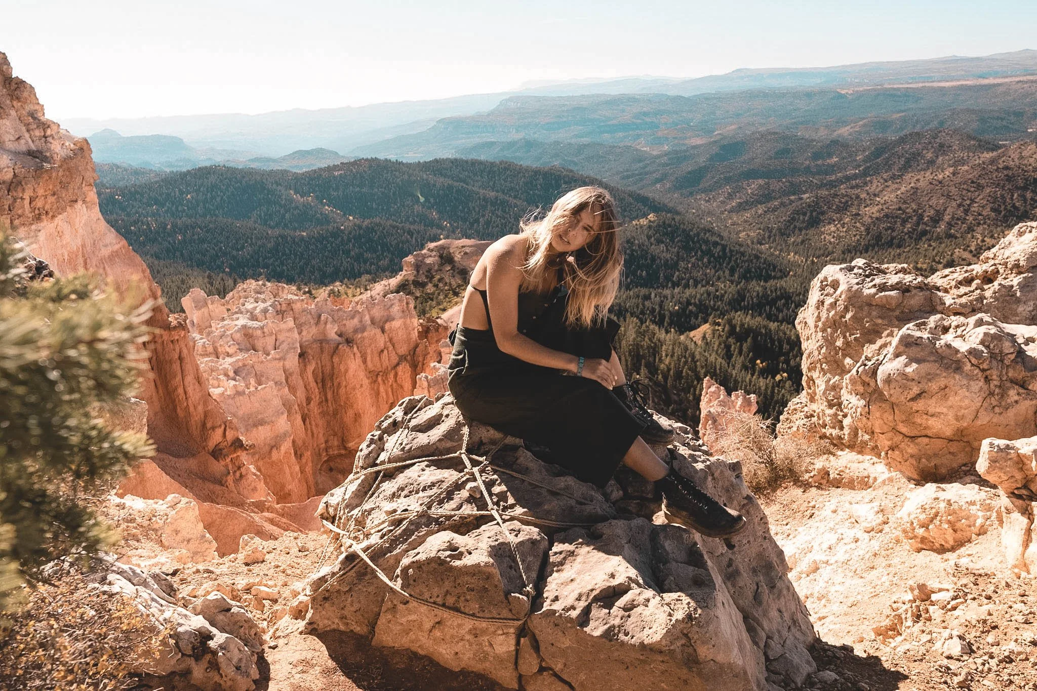 A young woman sitting on a rock formation on a mountain, with a view of forested hills and valleys in the background.