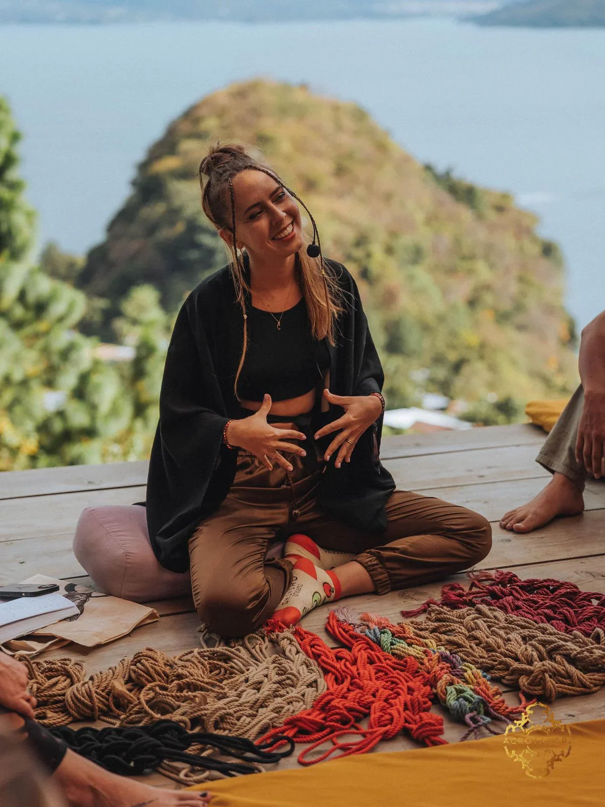 A woman sitting cross-legged on a wooden deck, smiling and speaking with a microphone headset, with colorful knitted scarves or shawls laid out in front of her, background of a mountain and water.