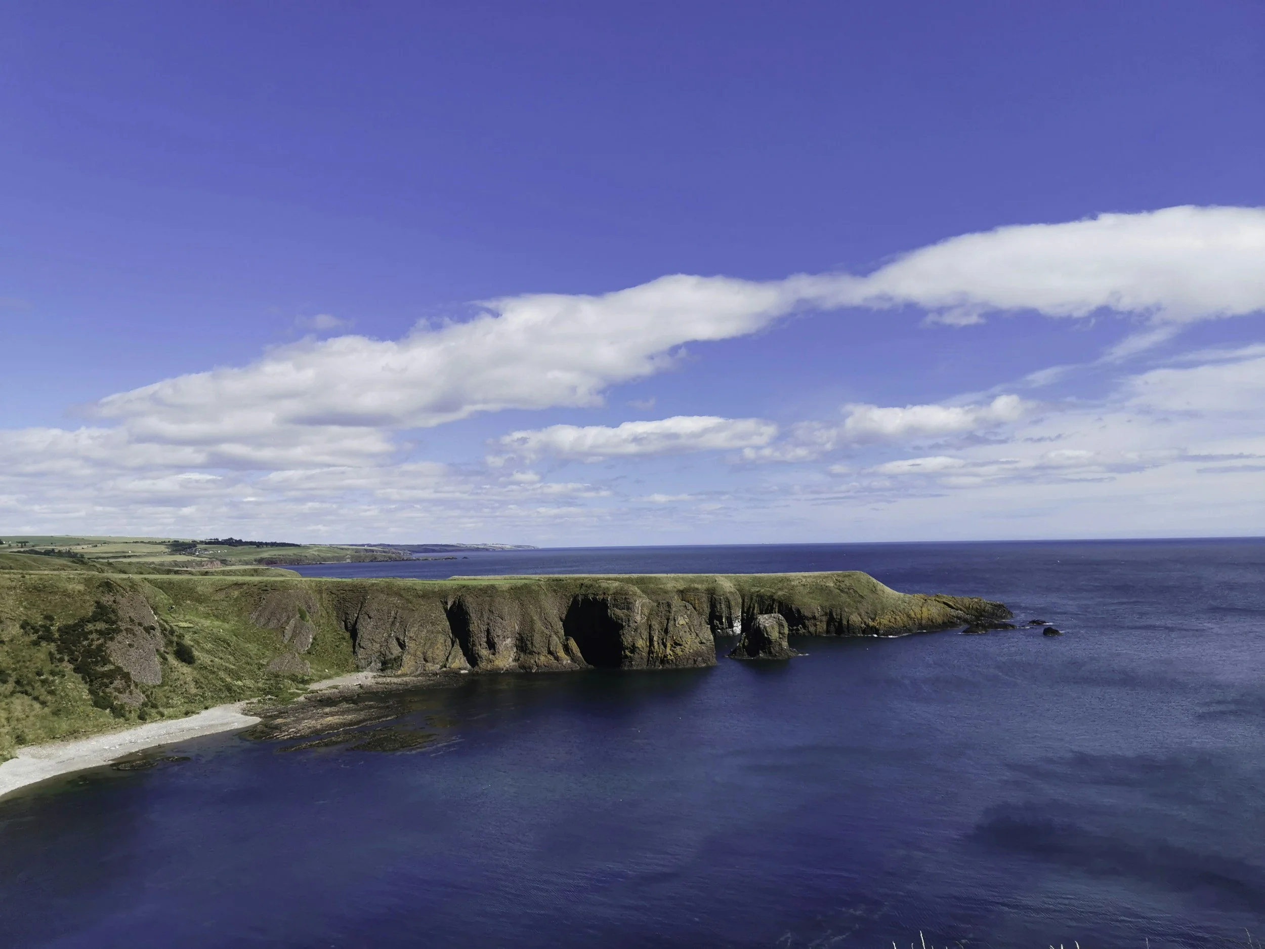 A coastal landscape with cliffs, green fields, and a beach, under a blue sky with scattered clouds.