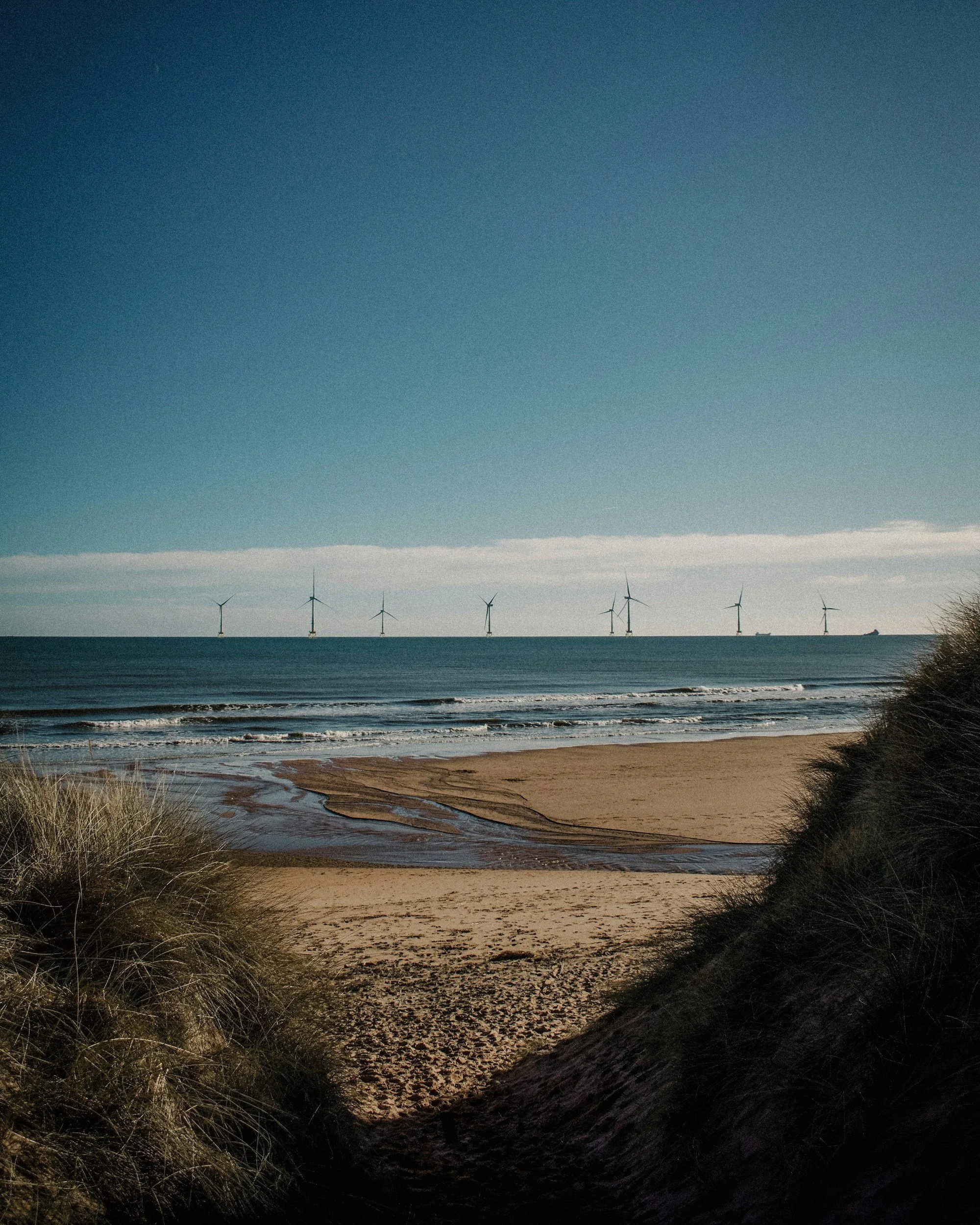 Beach with sandy shoreline, grassy dunes in the foreground, the ocean waves, and wind turbines in the distance under a clear sky.