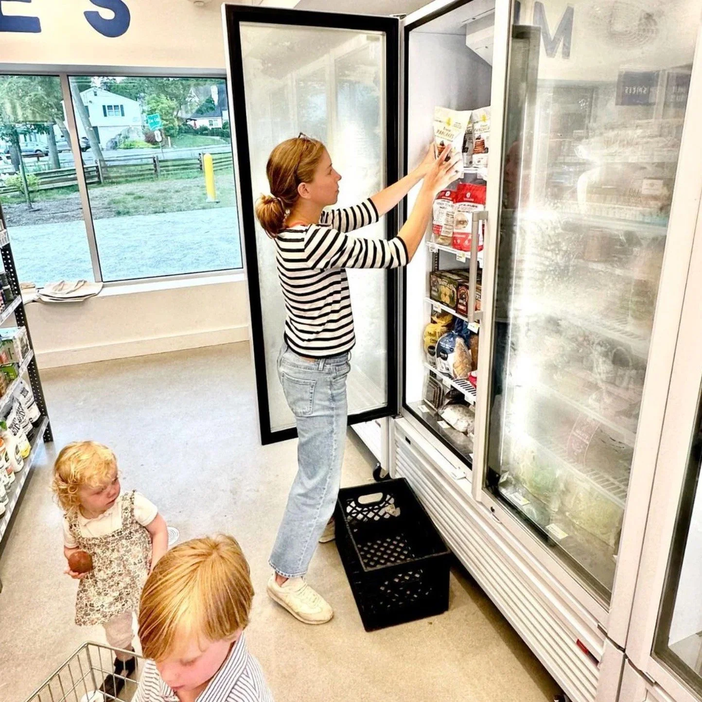 A woman in a striped shirt and jeans stocking a refrigerator in a grocery store while two children watch, one holding a chocolate snack.