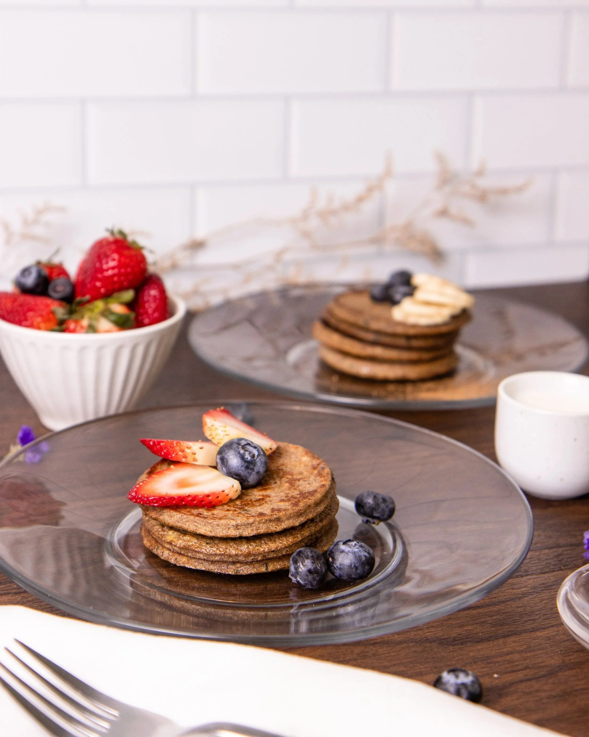 Plate of four chocolate pancakes topped with sliced strawberries and blueberries on a wooden table set for breakfast.