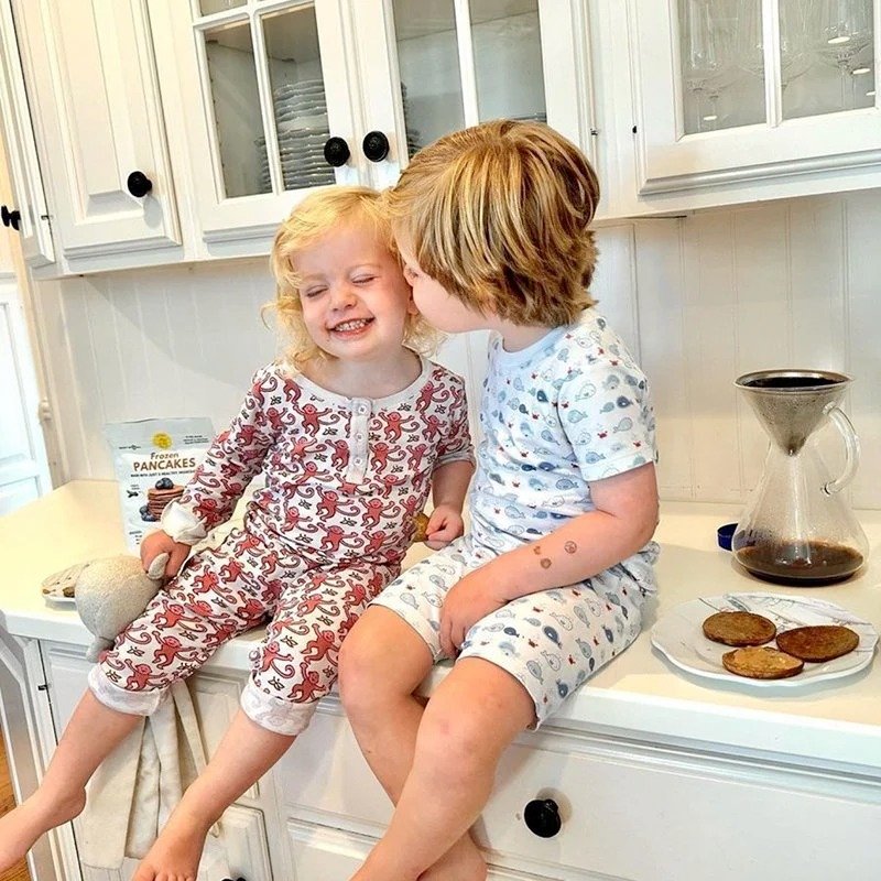 Two young children sitting on a kitchen counter, smiling and making a funny face at each other. The girl on the left is wearing a red and white patterned pajama set, and the boy on the right is in light blue pajamas with a pattern. There are pancakes and a coffee pot on the counter behind them.
