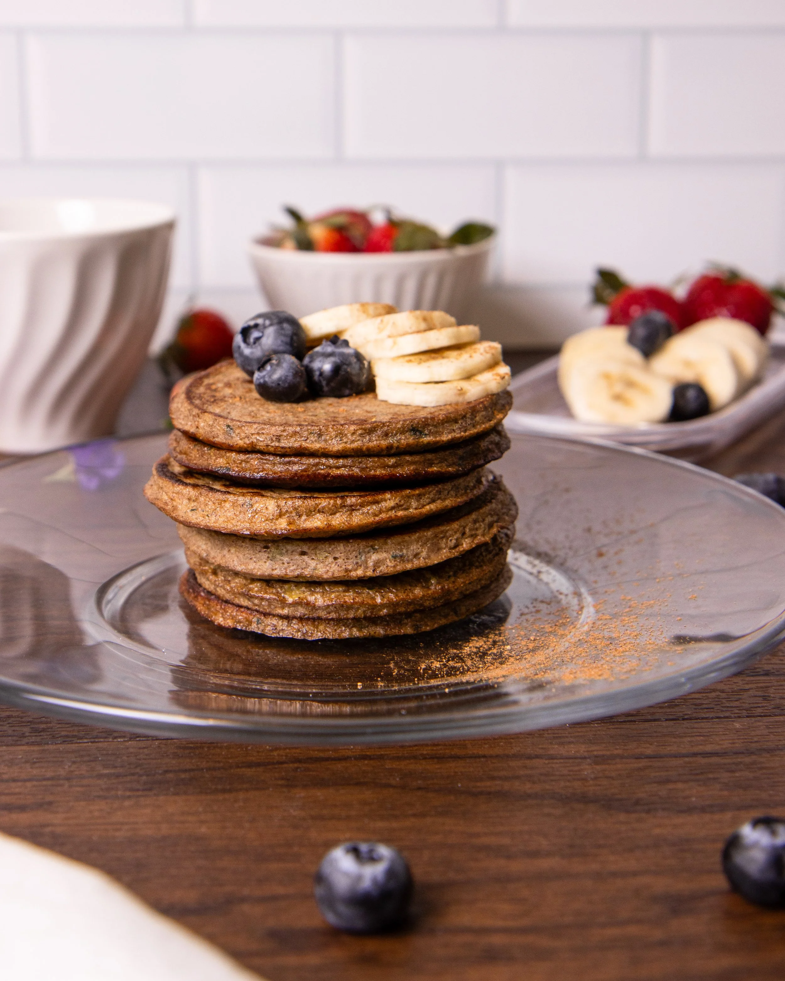 Stack of chocolate pancakes topped with sliced bananas and blueberries on a glass plate, with assorted berries and bowls in the background.